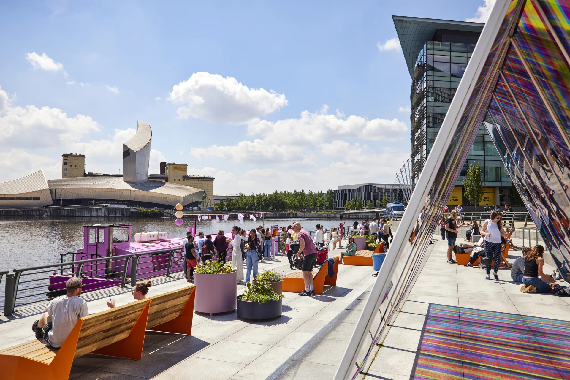 People relaxing near the water at MediaCityUK Quayside, with The Lowry building and a colorful art installation in the background.