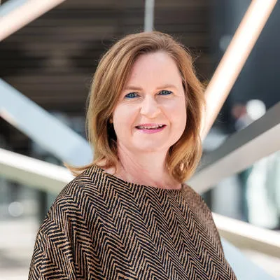 Woman with blonde hair smiling in a patterned top indoors with modern architecture.