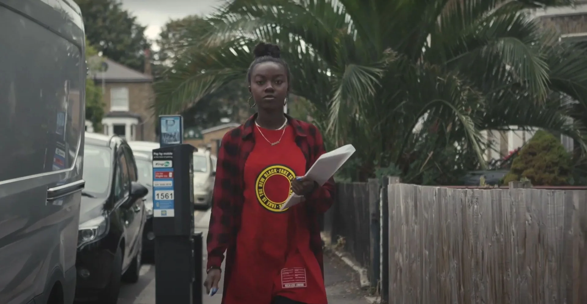 Young Black woman wearing a red branded shirt and plaid overshirt, carrying a document while walking down a suburban street next to parked vans and palm trees, promoting social value.