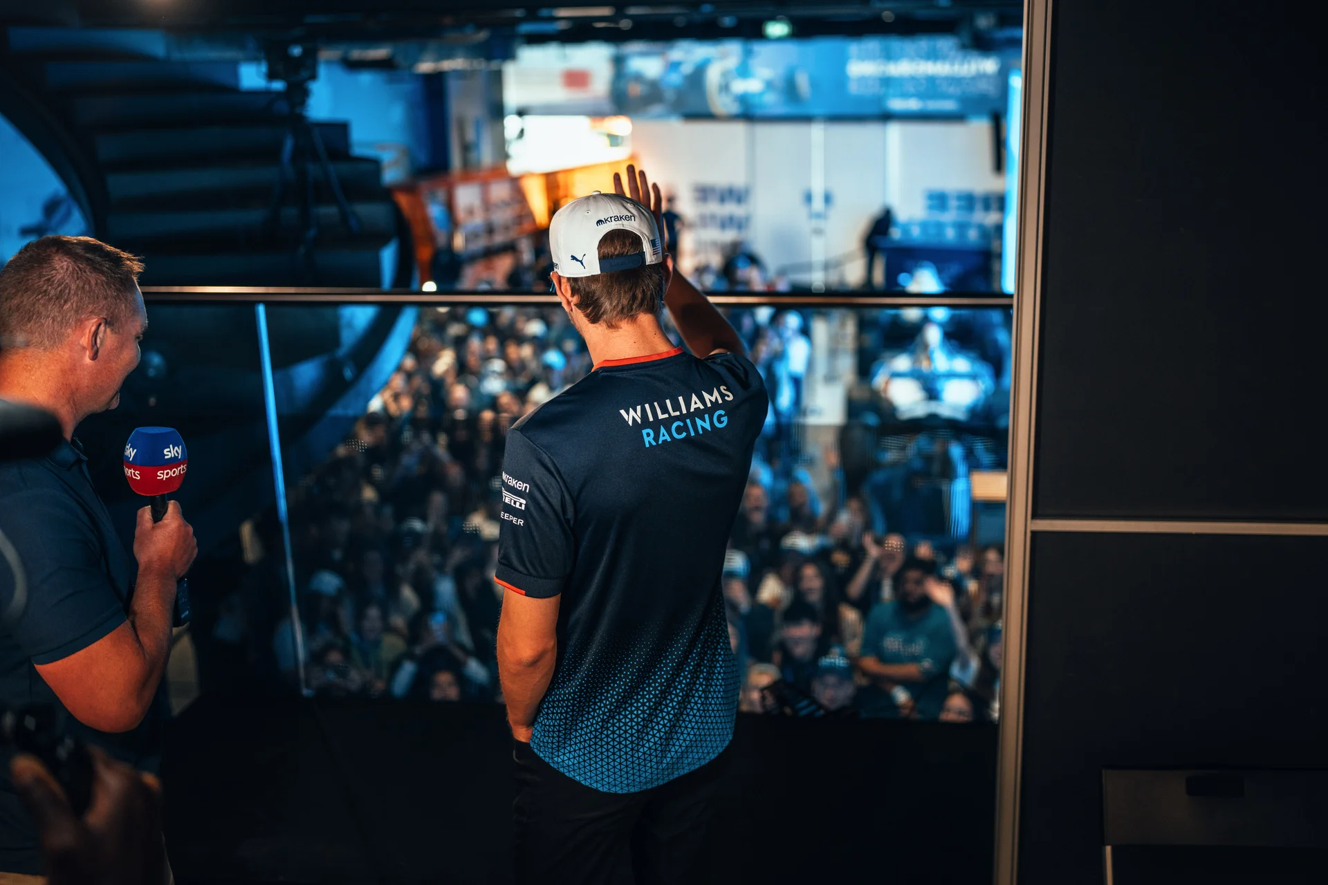 Williams Racing presenter waving to an audience during a live event, with a host interviewing nearby.