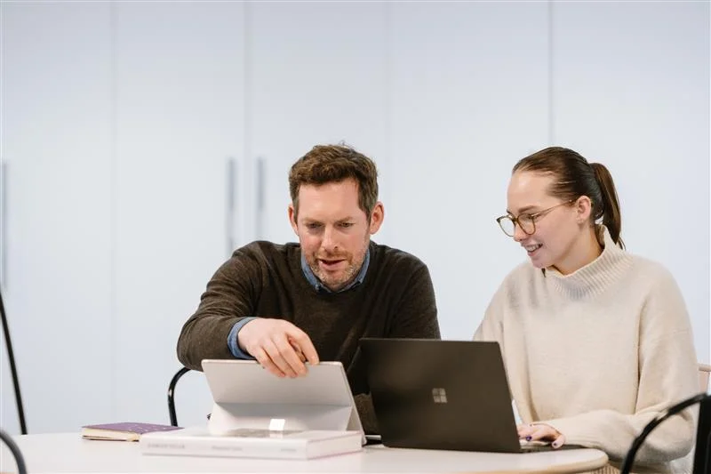 A man points at a tablet screen while talking to a smiling woman with glasses who is working on a laptop. They are sitting together at a table in a bright, modern office.