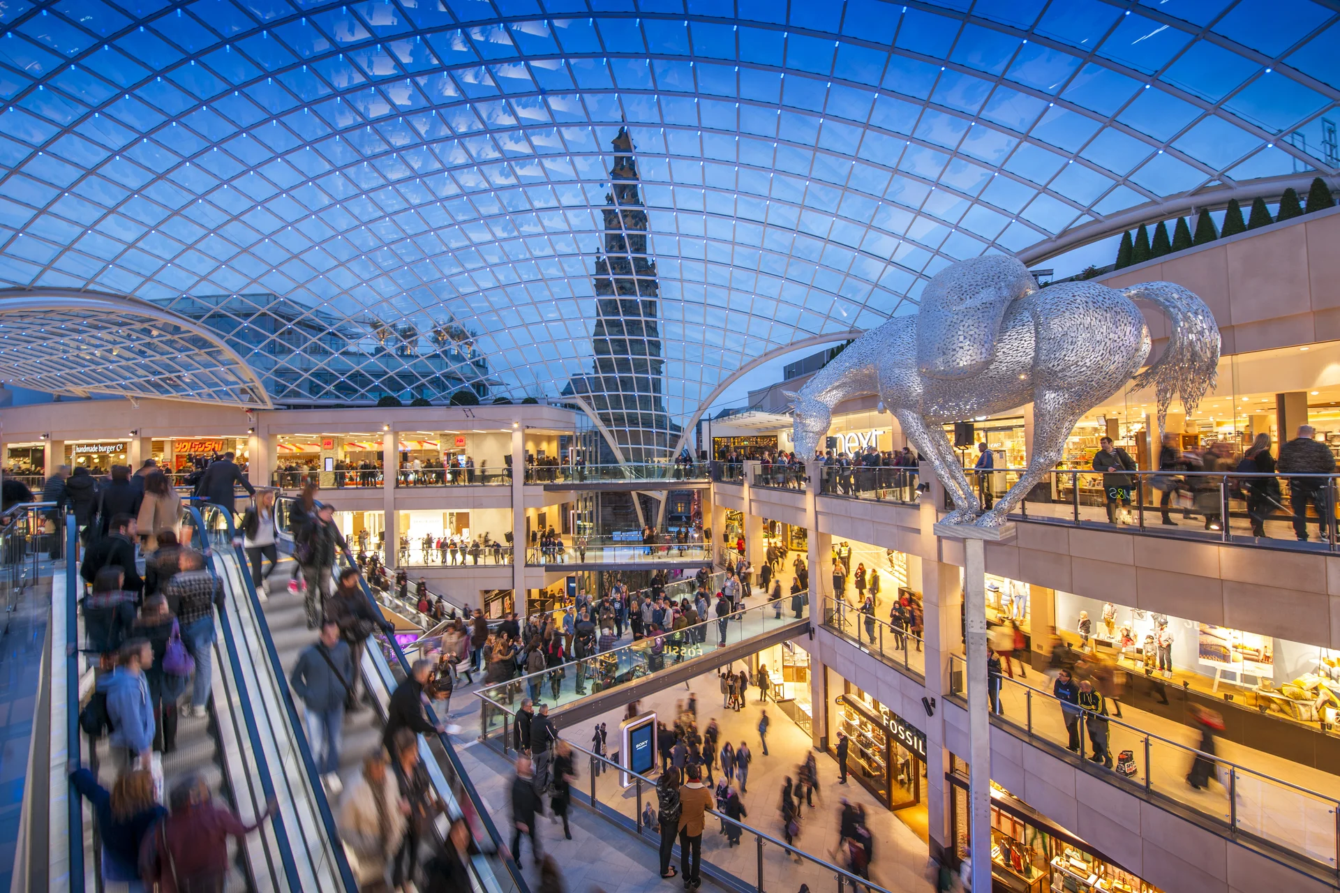 A bustling indoor shopping mall with a glass roof, escalators, and a large metallic lion sculpture, under a twilight sky.