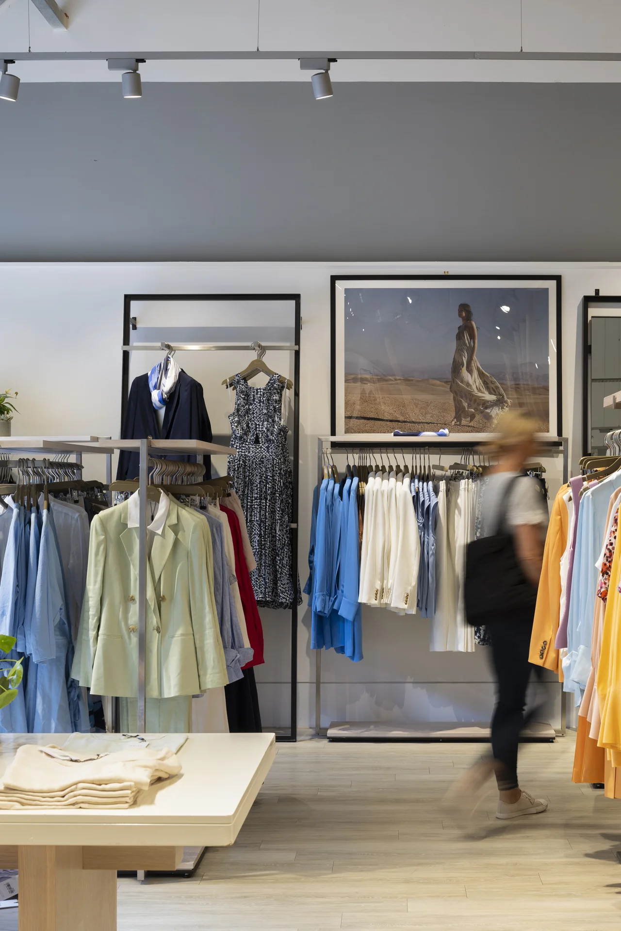 Interior of a fashion retail store at **Braintree Village** with racks of clothing in pastel and blue tones, a display table, and a motion-blurred shopper walking past a large framed advertisement.