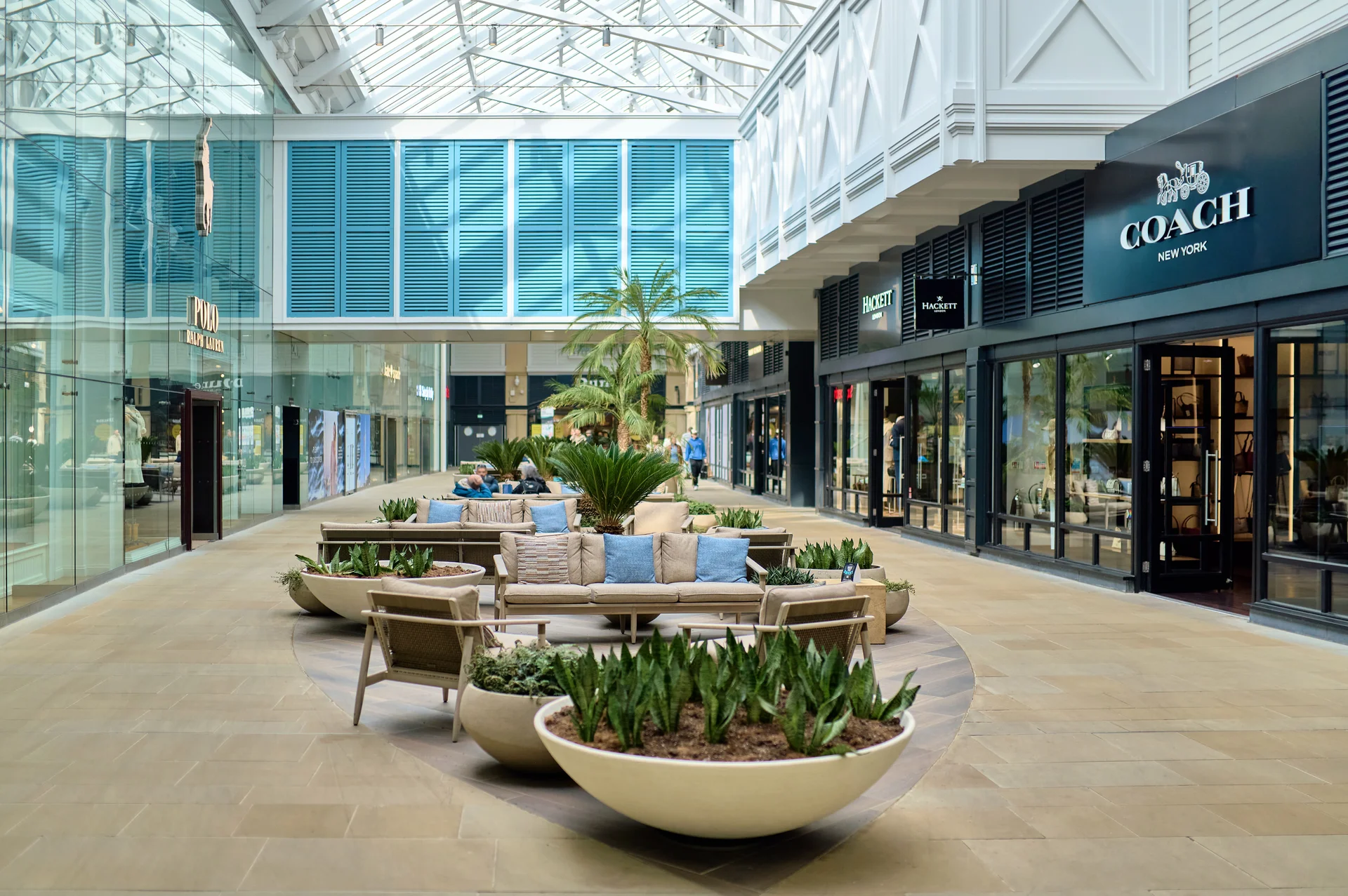 Interior of **Gunwharf Quays** shopping mall, featuring a central lounge seating area with planters under a skylight, surrounded by retail storefronts including **Coach** and **Hackett**.