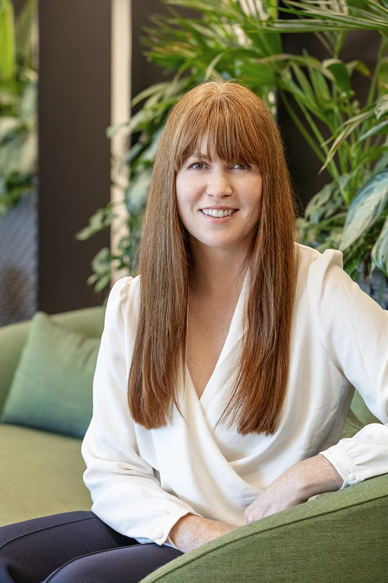 A professional portrait of Natasha Morris, Director of Flex Workplace. She is smiling warmly while seated on a sofa in a bright, modern office filled with lush greenery.