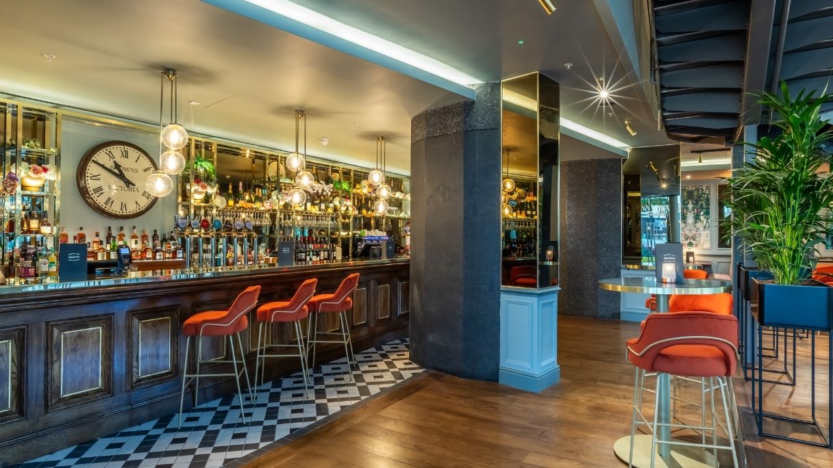 The bar at Browns with patterened floor, red velvet stools, and a large clock next to the bar stock