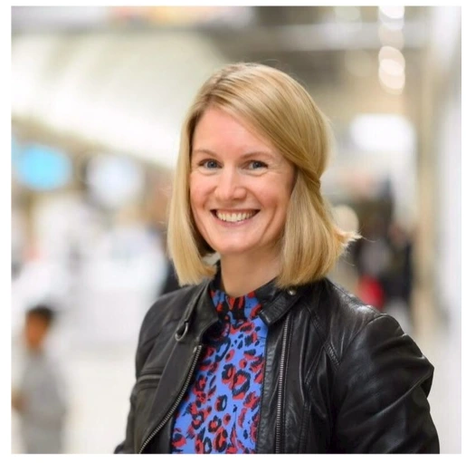 Blonde woman in black leather jacket and patterned shirt smiling.