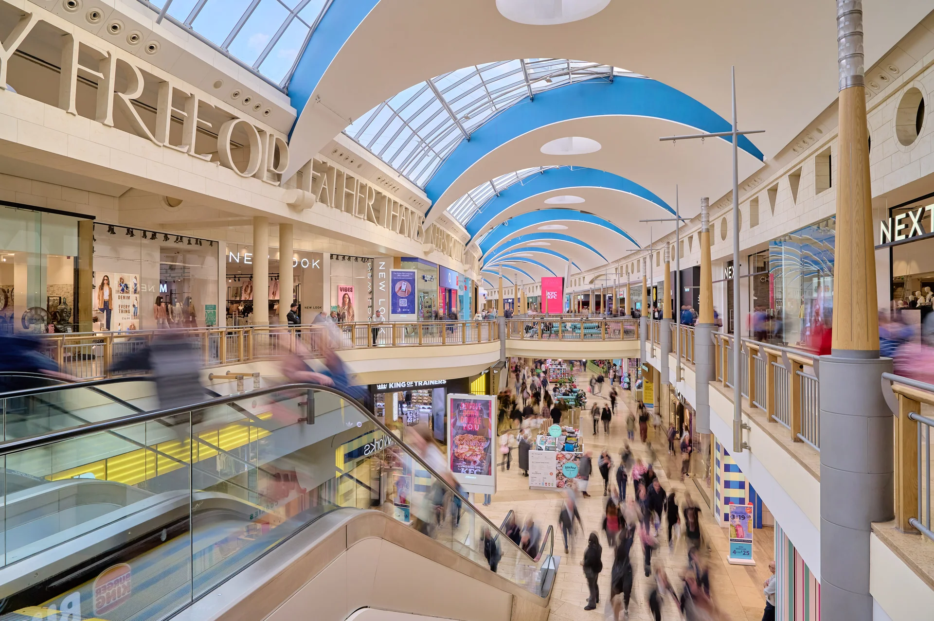 Wide-angle interior view of a busy atrium or gallery inside **Bluewater Shopping Centre**, showing multiple levels, escalators with motion blur, overhead skylights, and retail stores like NEXT and New Look.