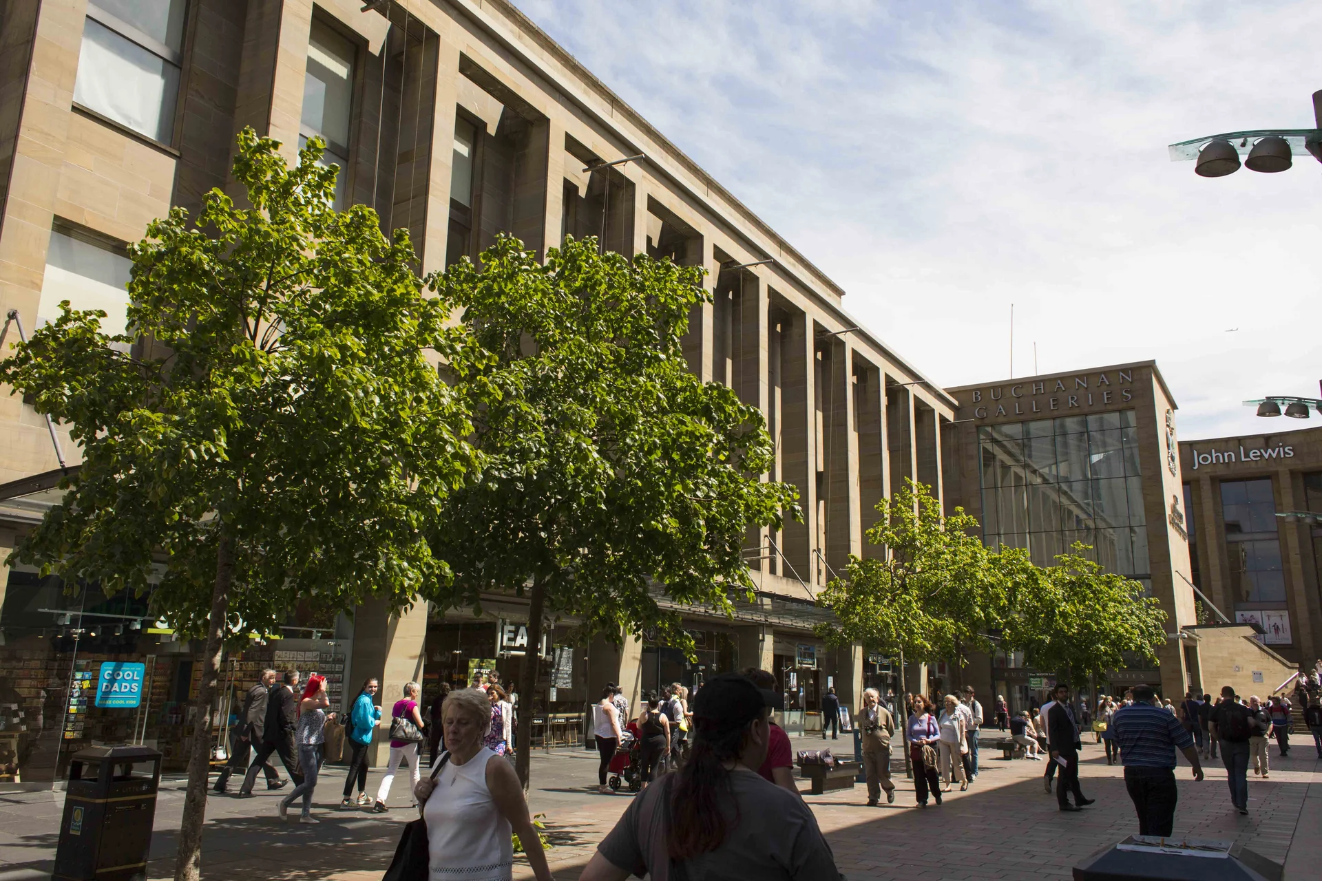 Exterior view of **Buchanan Galleries** in **Glasgow**, emphasizing the long, columnar stone facade and the street-level retail units shaded by green trees, with pedestrians walking in the public realm.