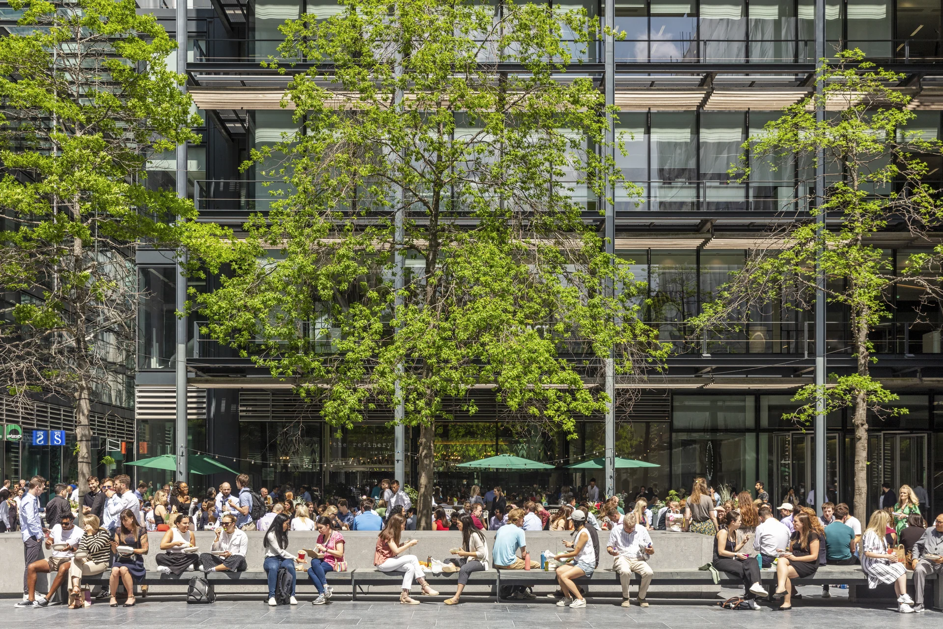 Busy office plaza with people sitting on benches, green trees, and a glass building in the background