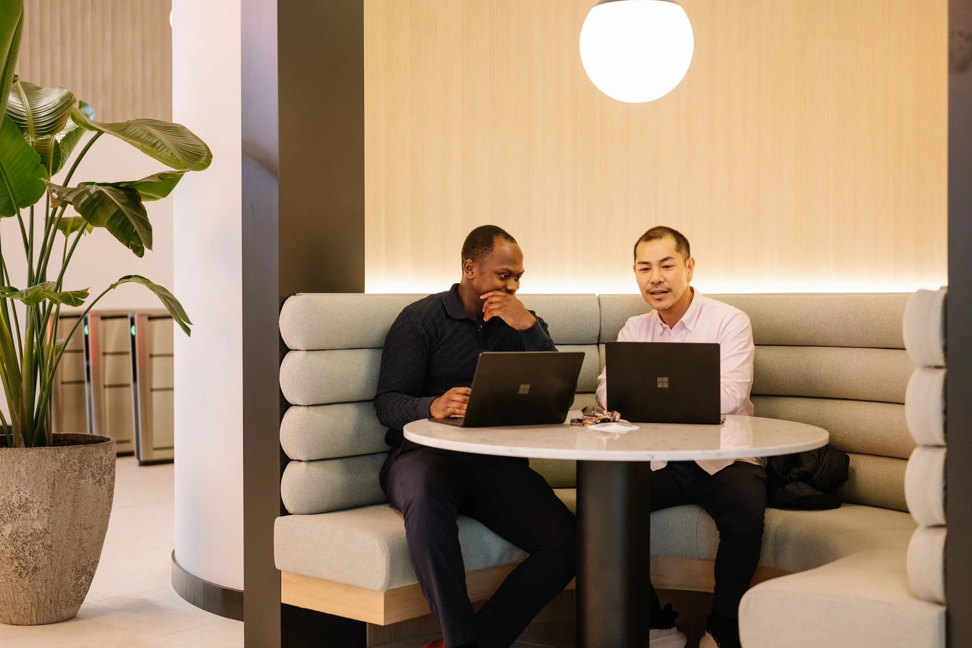 Two men seated on a cushioned bench, working on laptops at a round table in a modern, well-lit interior space.