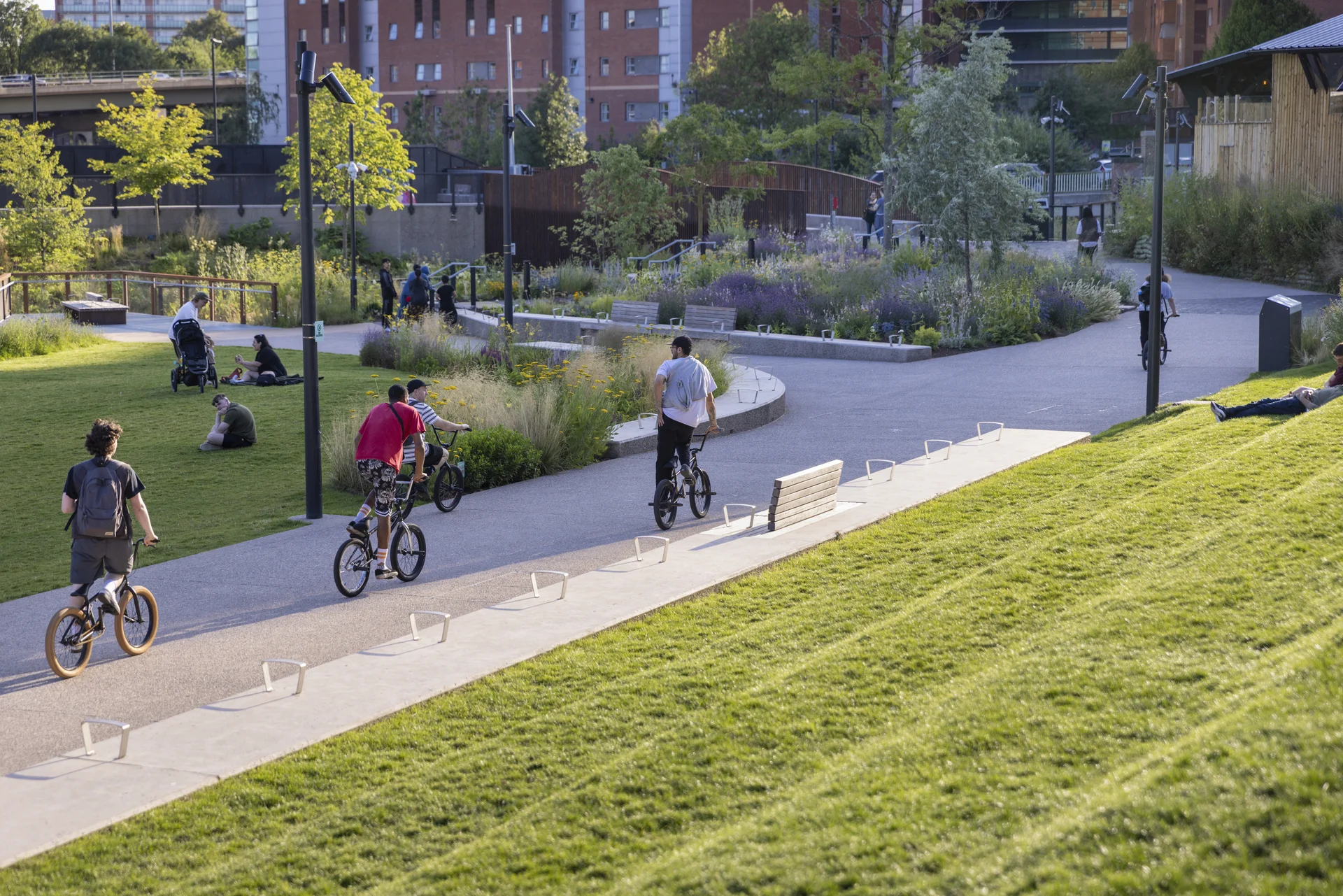 Cyclists riding BMX bikes on a paved path through **Mayfield Park** in Manchester, showing large grassy hills and landscaped beds on a sunny day.