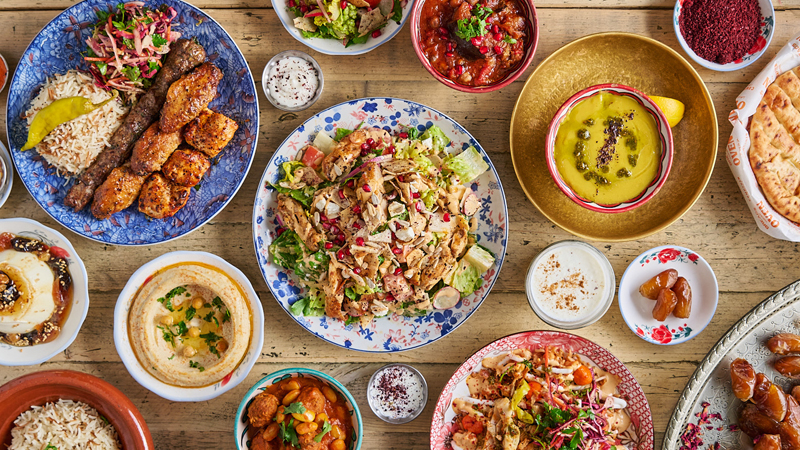 A shot from above of a beautiful feast on a table with colourful various sized plates and a vaiety of lebanese dishes such as hummus, flat bread, grilled meat, salad, yoghurt and more