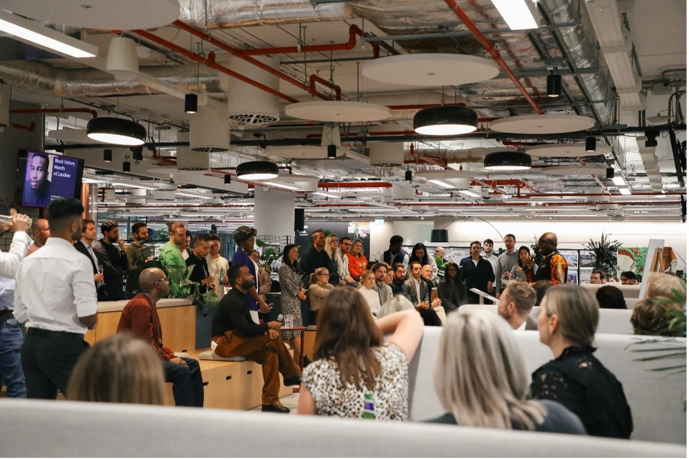 A diverse group of people gathered in a modern, open-plan office space with exposed ceilings and industrial lighting.