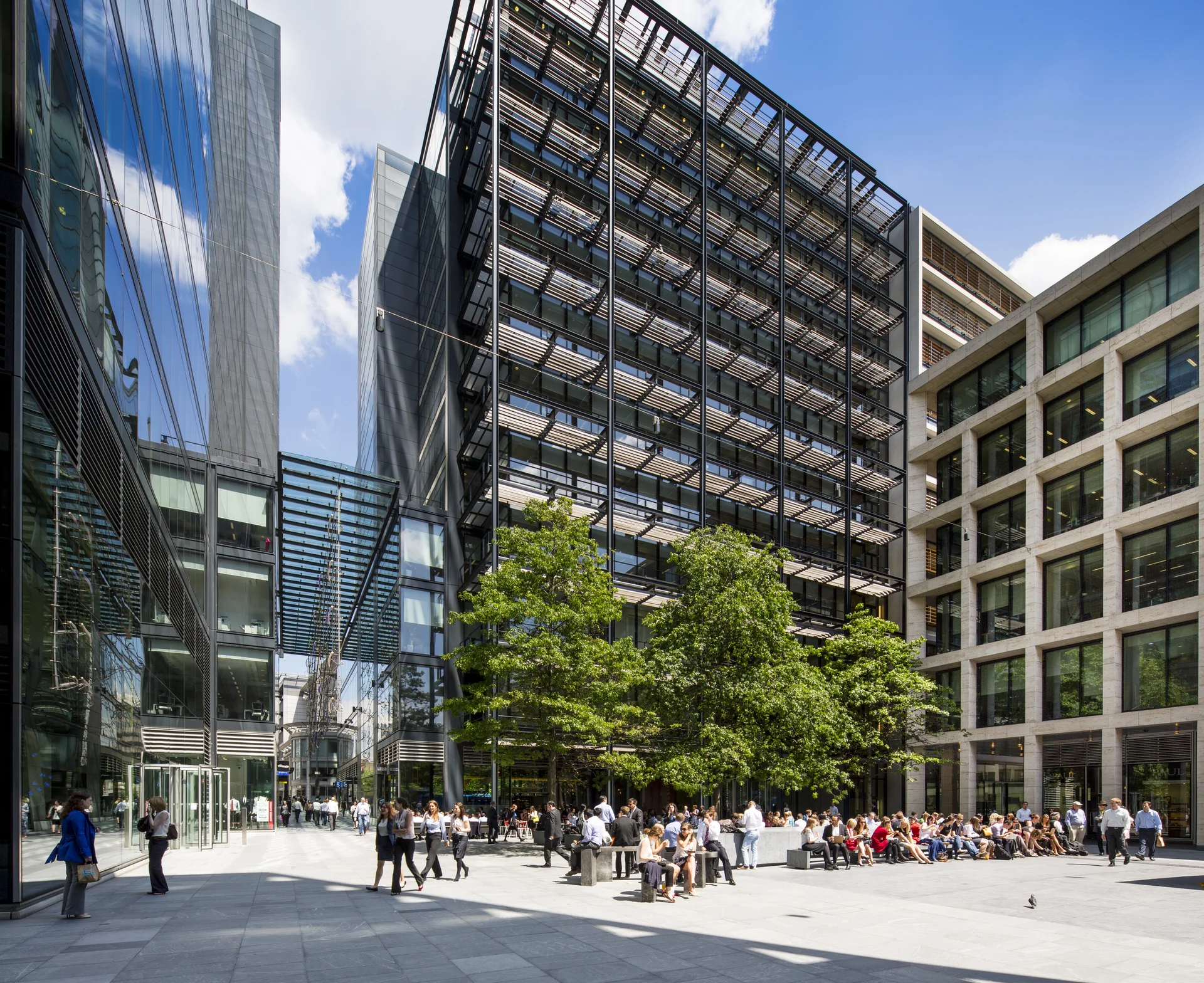 Modern glass and steel buildings surround a plaza with people sitting and walking, trees providing shade, under a clear blue sky.