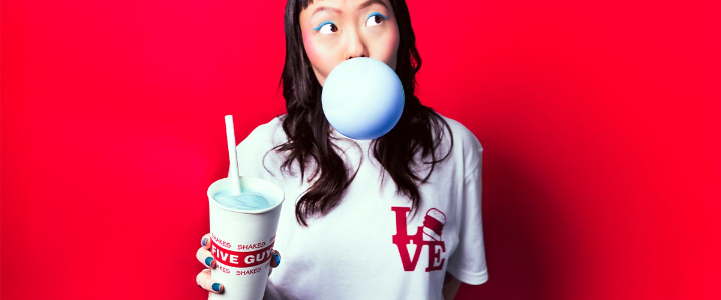 A girl with blue eyeliner on blowing a blue bubble and holding a Five Guys milkshake