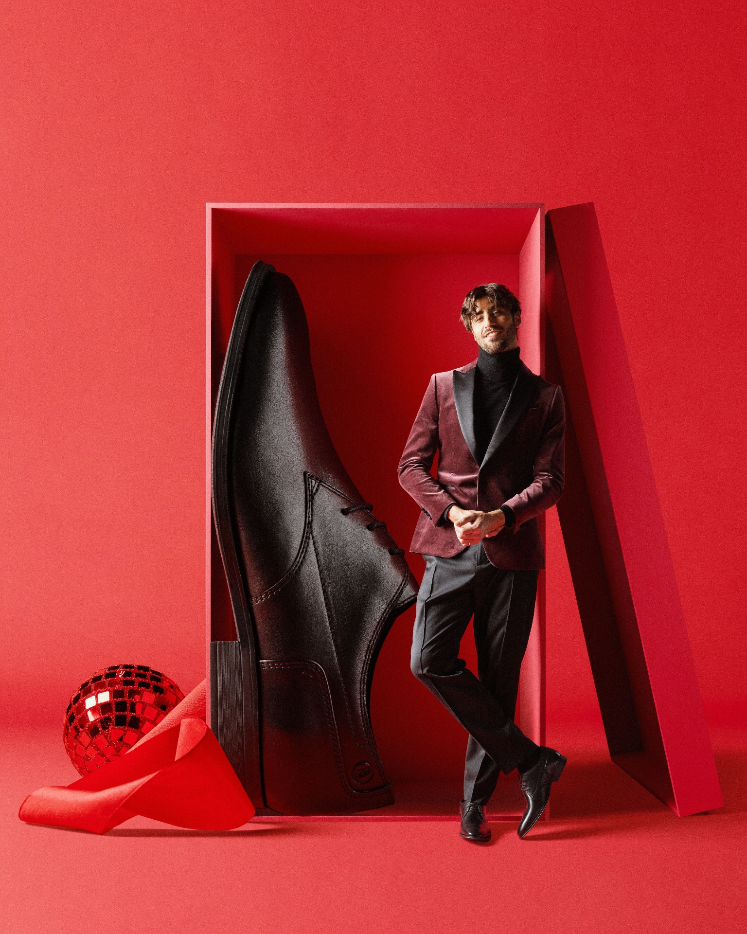 A man in a red blazer leans against a giant red shoebox 