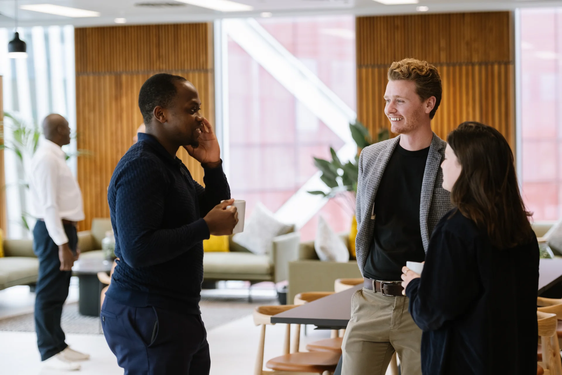 Three people conversing in a modern, well-lit office space with wooden walls, comfortable seating, and large windows.