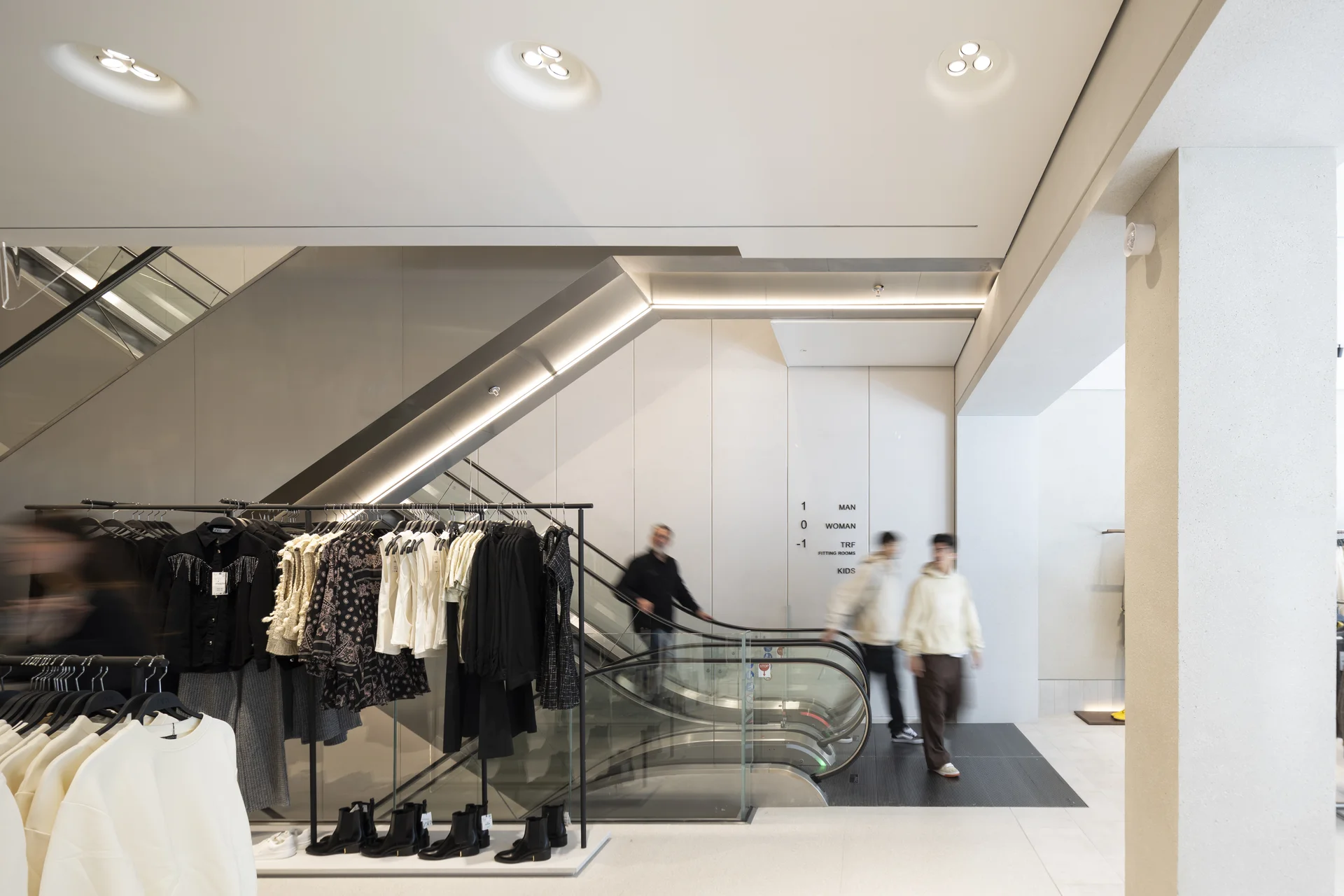 Minimalist interior of a retail store (Zara) in Trinity Leeds. Clothing racks and escalator with floor directions.