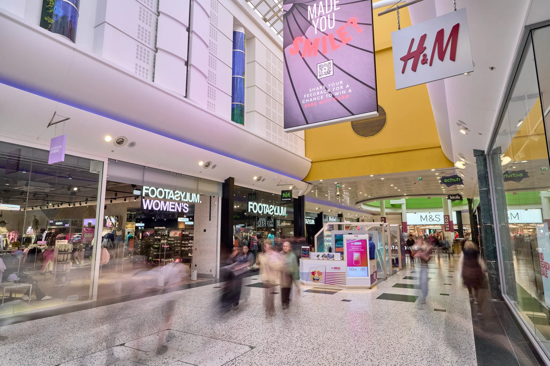 Wide interior view of the **White Rose Shopping Centre** mall, highlighting the storefronts for **FOOTASYLUM Women's** and **H&M**, with a large overhead digital advertisement and shoppers moving through the corridor.