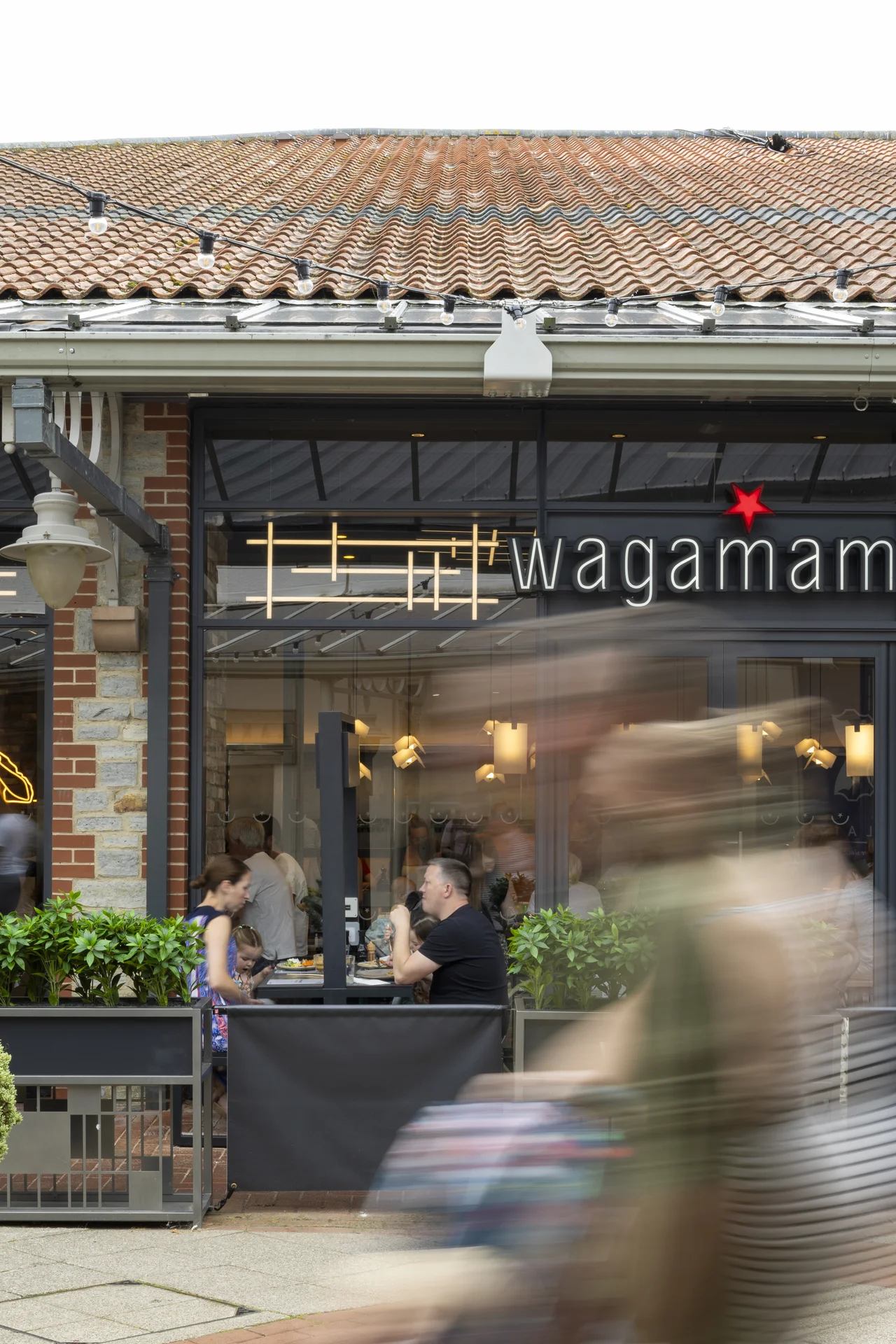 Close-up of the **Wagamama** restaurant at **Clarks Village**, showing customers seated in the outdoor dining area with plants and a brick facade, viewed with motion-blurred shoppers passing in the foreground.