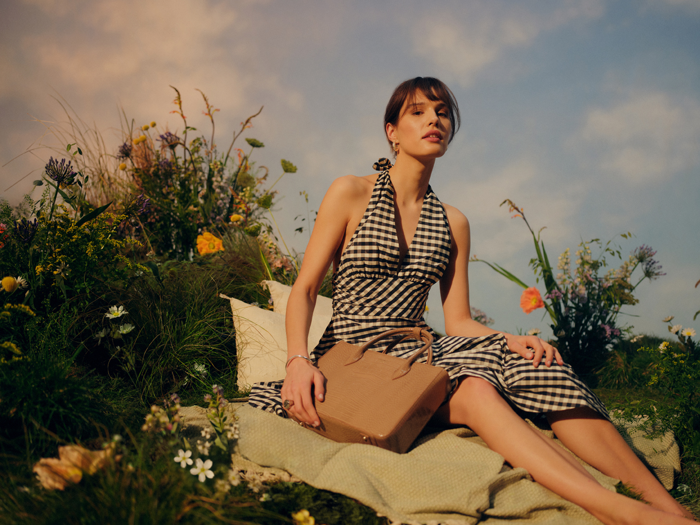 woman wearing a black and white gingham sundress sitting on a picnic blanket with a brown Osprey London bag. Flowers and grass can be seen against a superficial blue sky.
