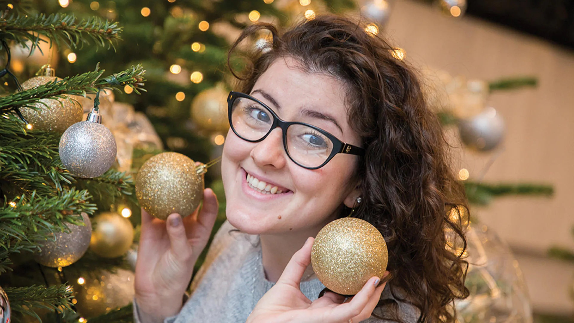 Person with curly hair and glasses holding golden Christmas ornaments in front of a decorated Christmas tree.