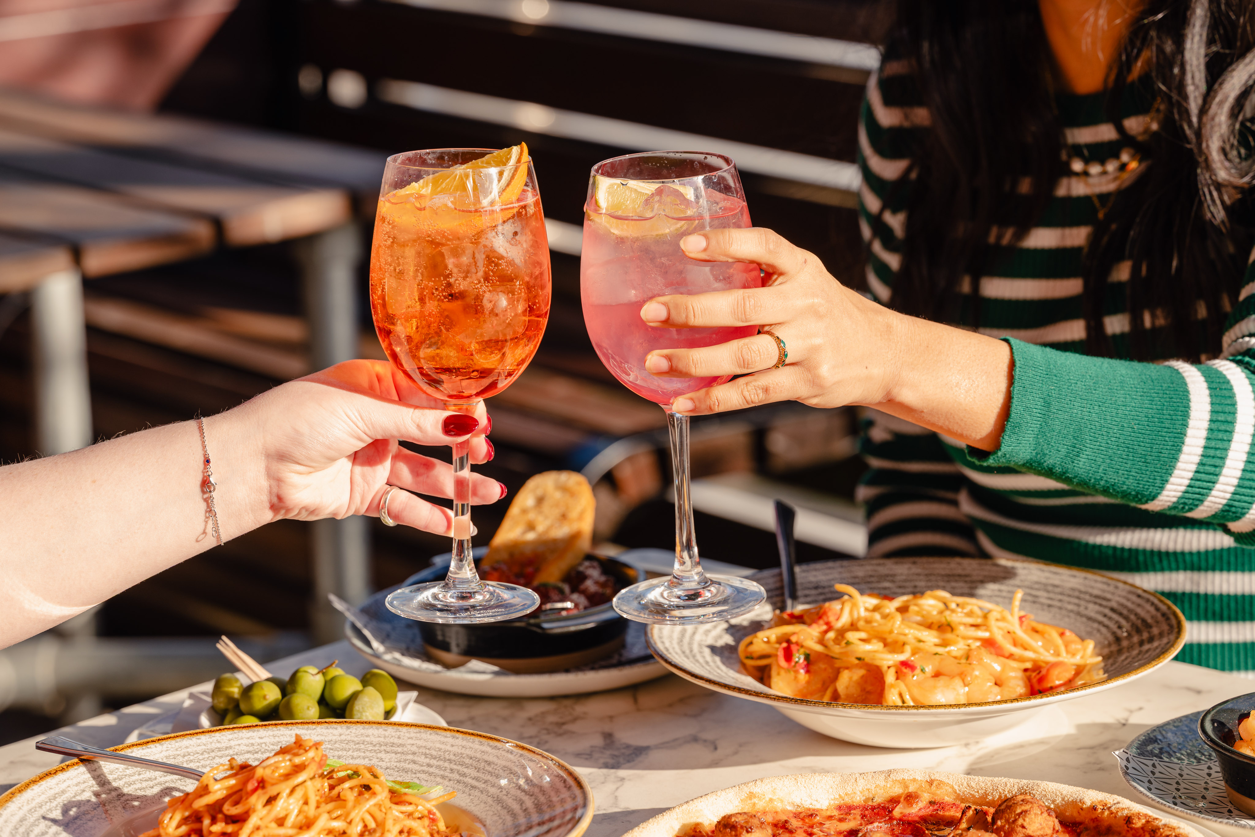 Two people clinking glasses over a table with spaghetti and other Italian dishes. The glasses contain a Spritz style cocktail.