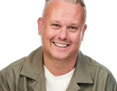 A man with short blond hair, wearing a green jacket over a white shirt, smiling against a white background.