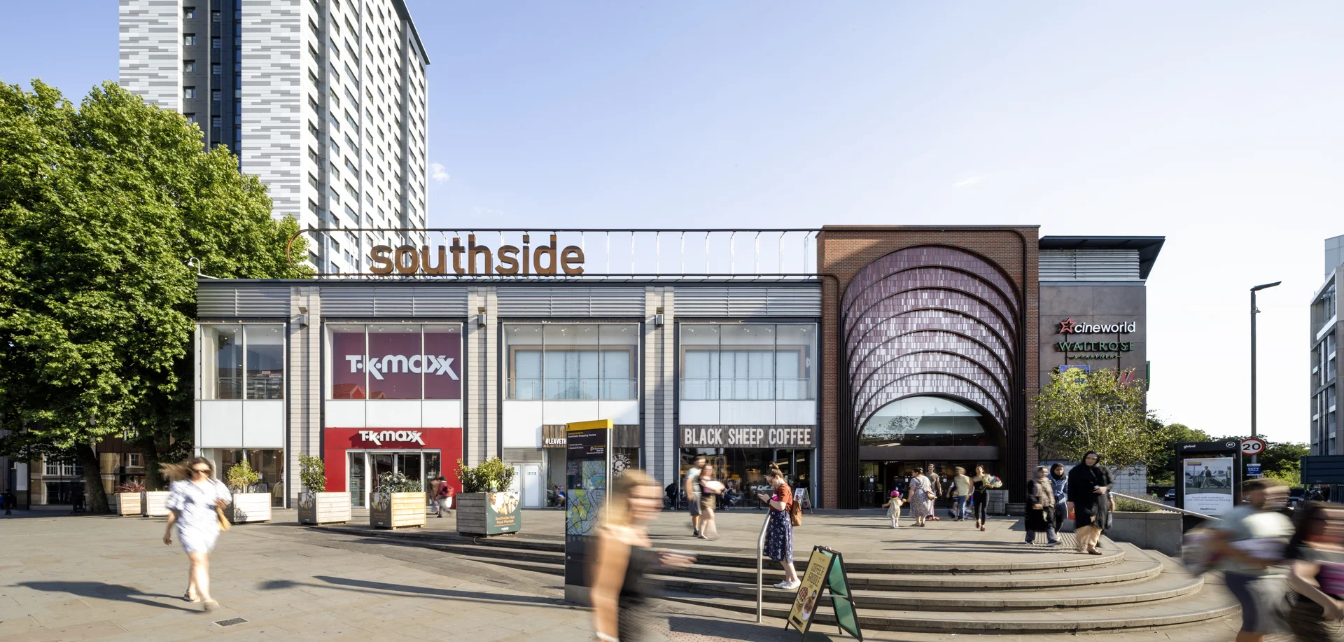 A modern shopping center with glass facades, a curved entrance, and people walking outside on a sunny day.