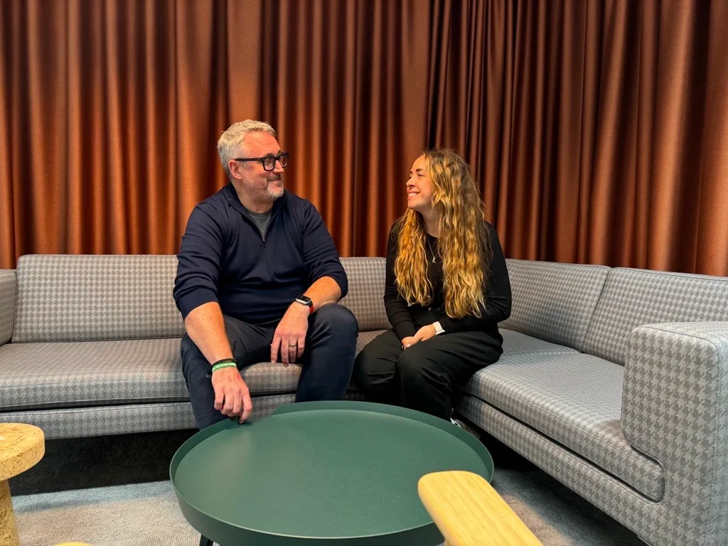 Two individuals sit on a patterned sofa, engaged in conversation, with a green table and brown curtains in the background.