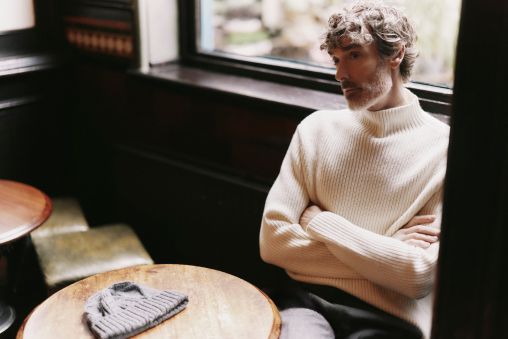 A man wearing a cream polo neck jumper sitting at a round wooden table