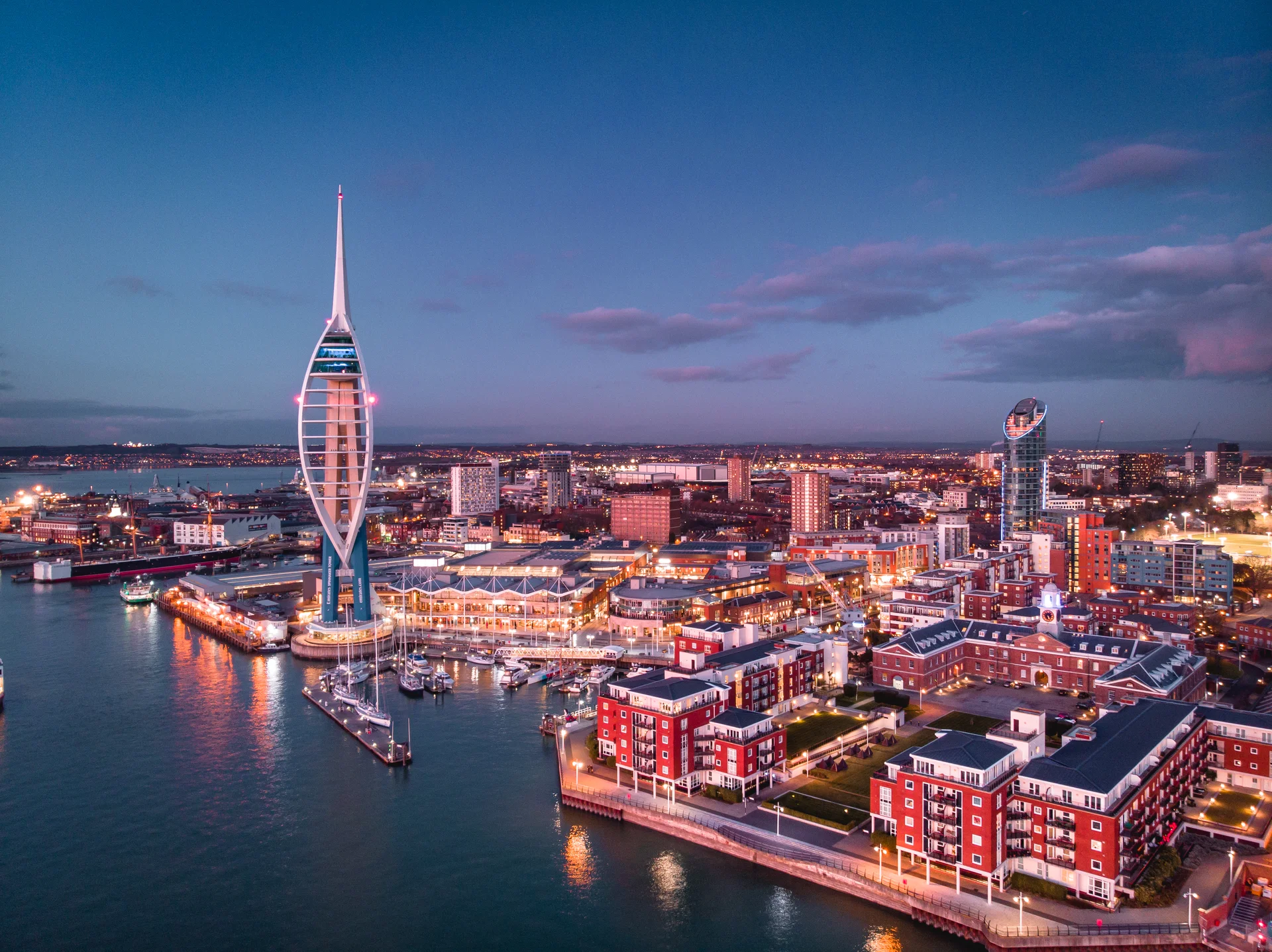 Evening cityscape with a prominent spire, illuminated buildings, waterfront, and boats, under a twilight sky.