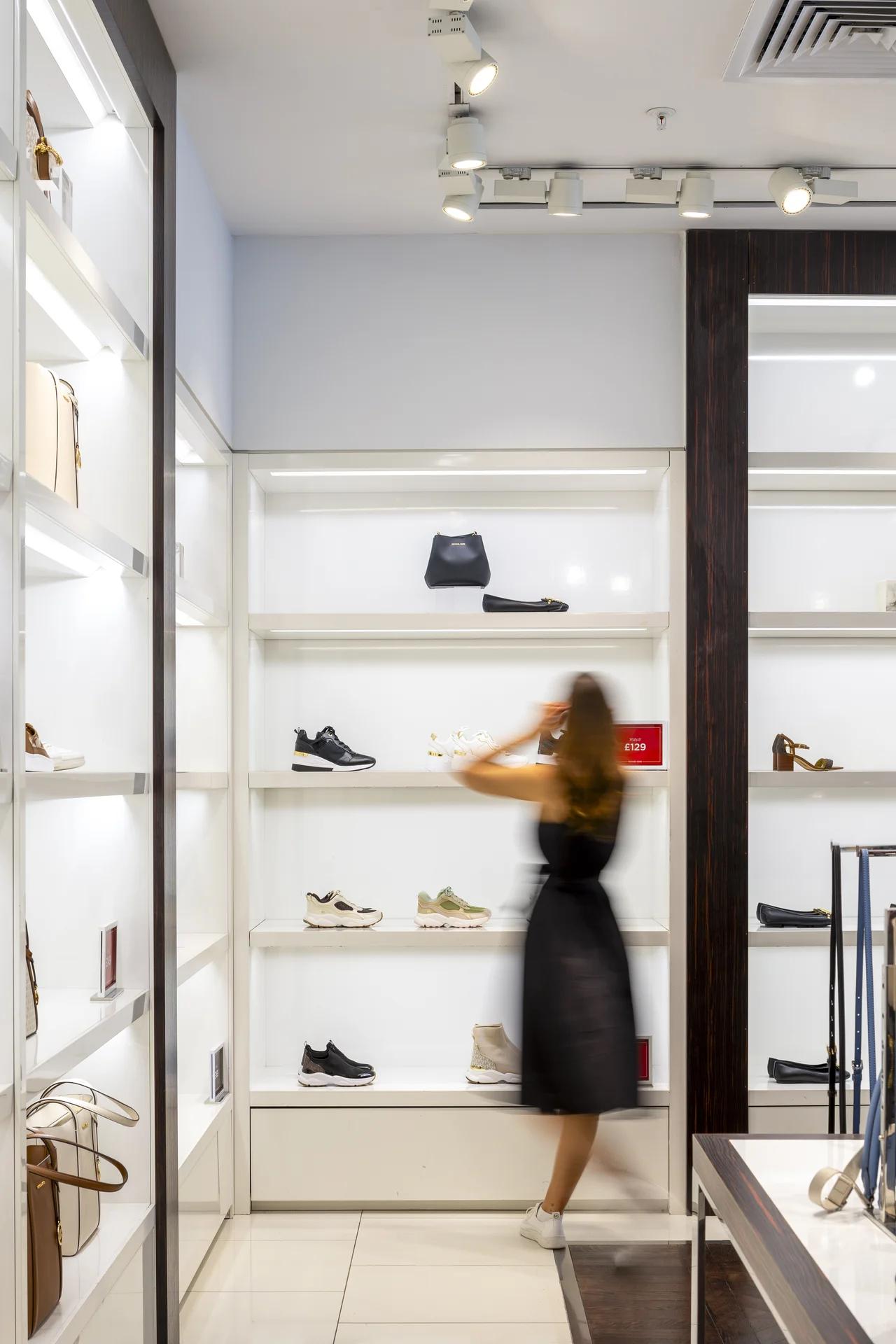 Bright, high-end retail interior in a **Gunwharf Quays** outlet store, showcasing brightly lit shelves displaying shoes and bags, with a motion-blurred shopper in a black dress browsing the merchandise.