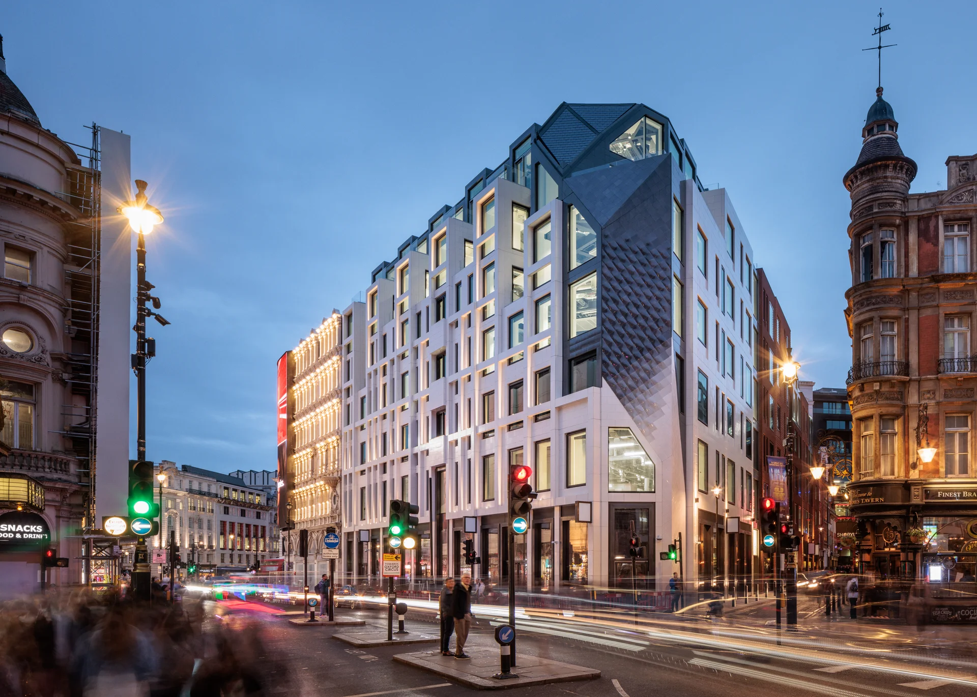 Modern building with geometric facade and illuminated windows at dusk, surrounded by historic architecture and busy street.