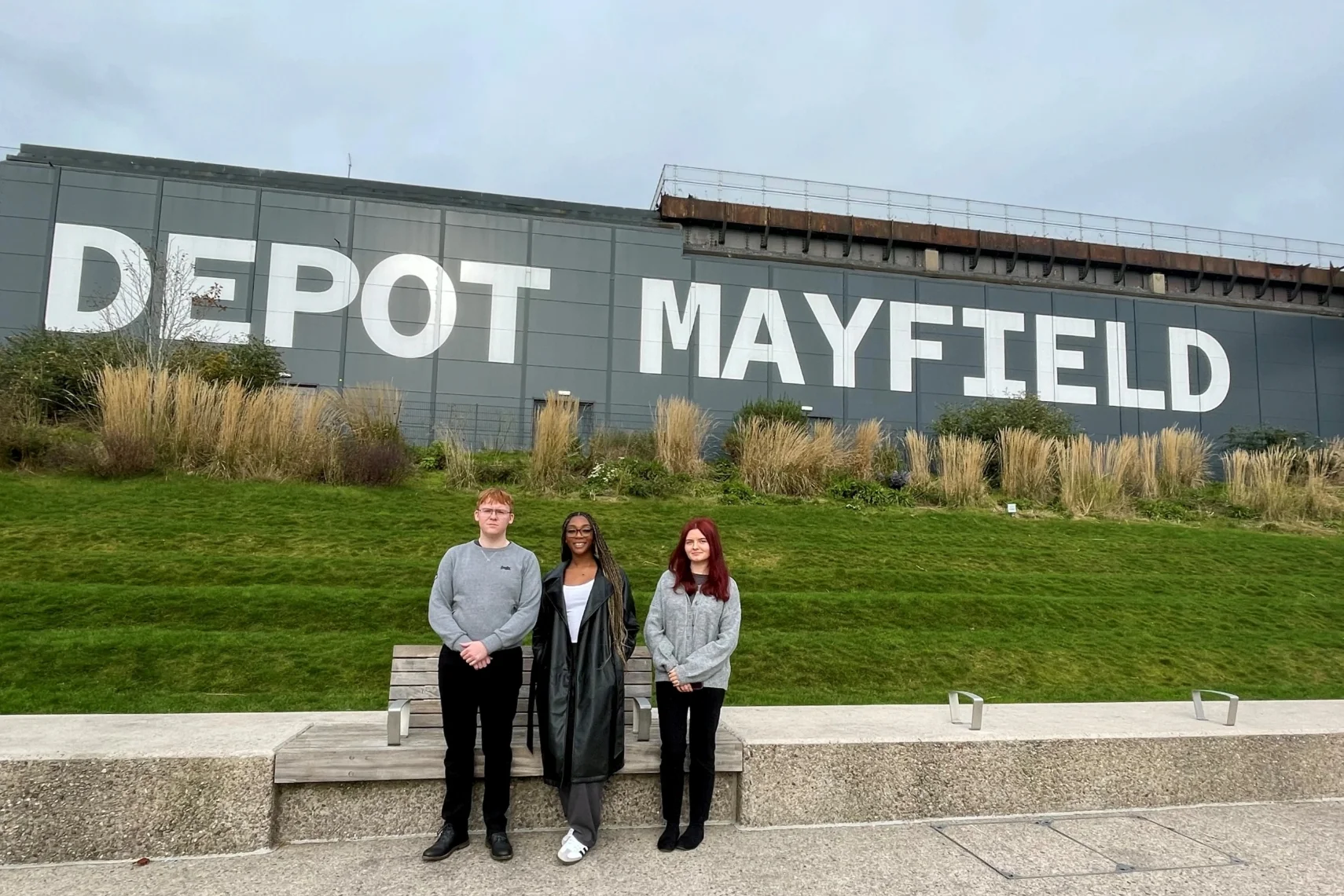 Three people stand in front of a grassy slope with 'DEPOT MAYFIELD' on a large wall, under an overcast sky.