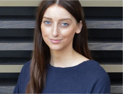 A person with long brown hair wearing a dark blue top, standing in front of a striped wooden background.