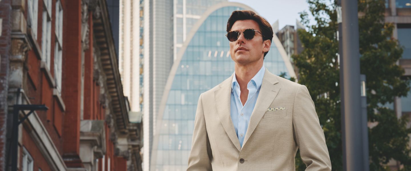 A male model wearing a cream suit on a city street in front of a glass building