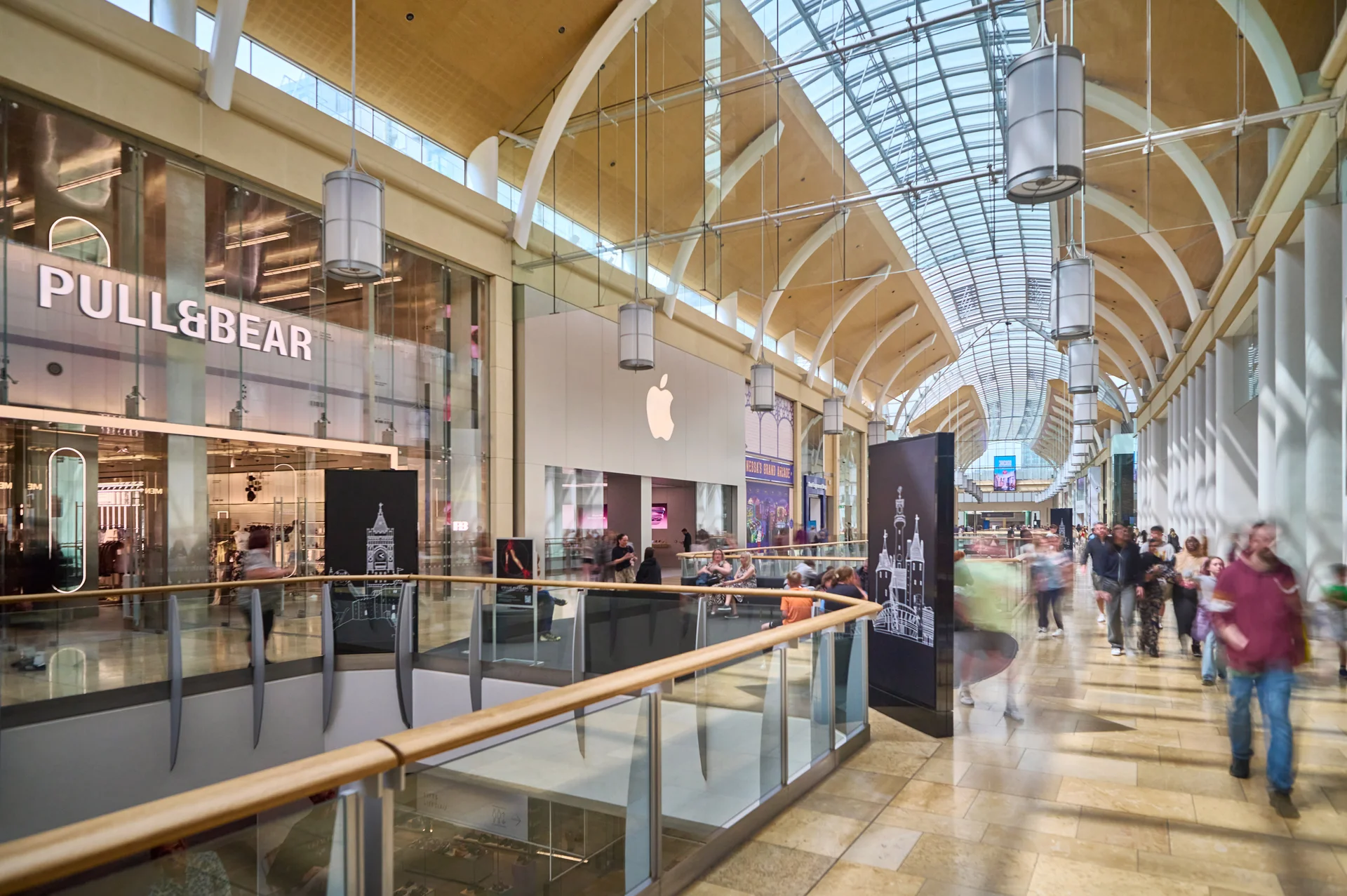 A modern shopping mall with arched ceilings, glass railings, and shoppers walking near a Pull&Bear store and an Apple logo.