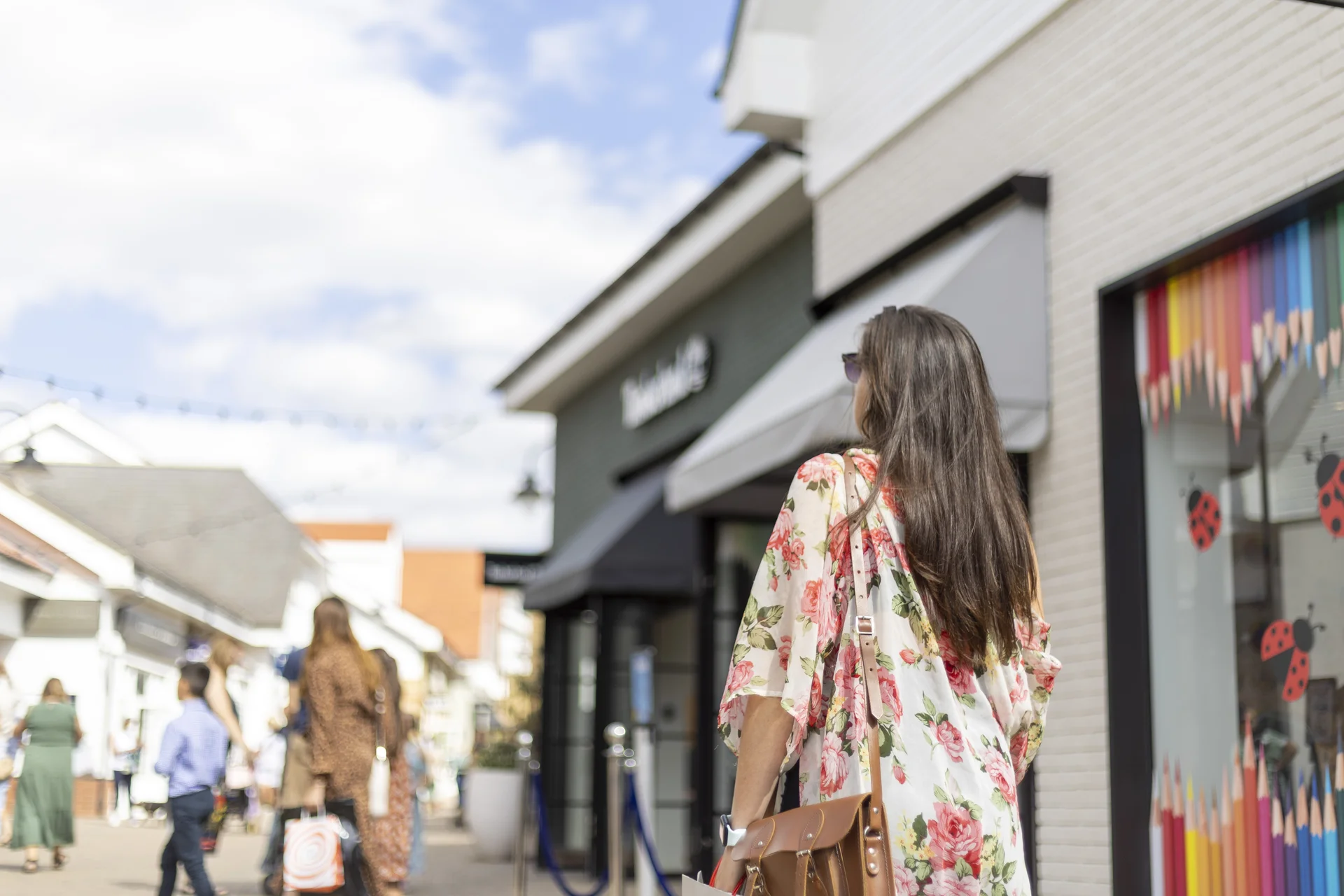 A shopper wearing a floral kimono and carrying a leather bag walks down the outdoor retail street at **Braintree Village**, with storefronts and other shoppers blurred in the background.