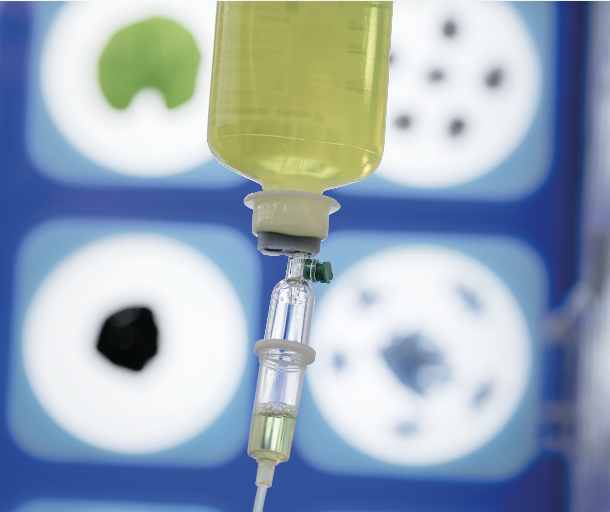 A close up image of an IV drip in a medical setting. The IV bag is green and there's a blurred blue background.