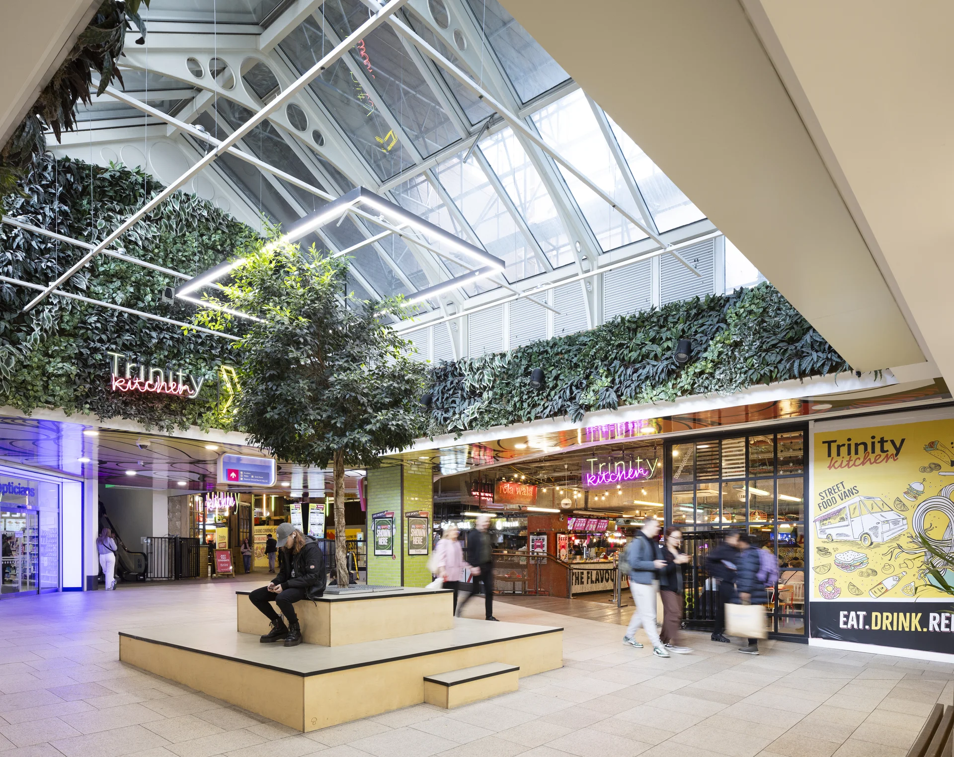 Trinity Kitchen area in Leeds, decorated with living walls, neon signs, and a central seating/planter area.