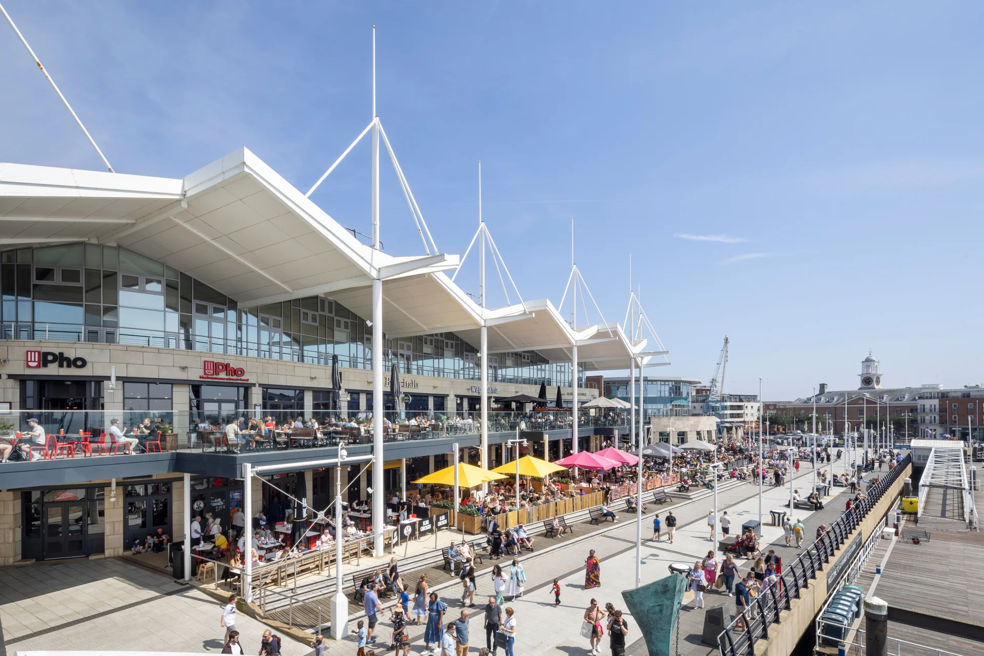Modern waterfront complex with glass facades, outdoor seating, colorful umbrellas, and people enjoying a sunny day.