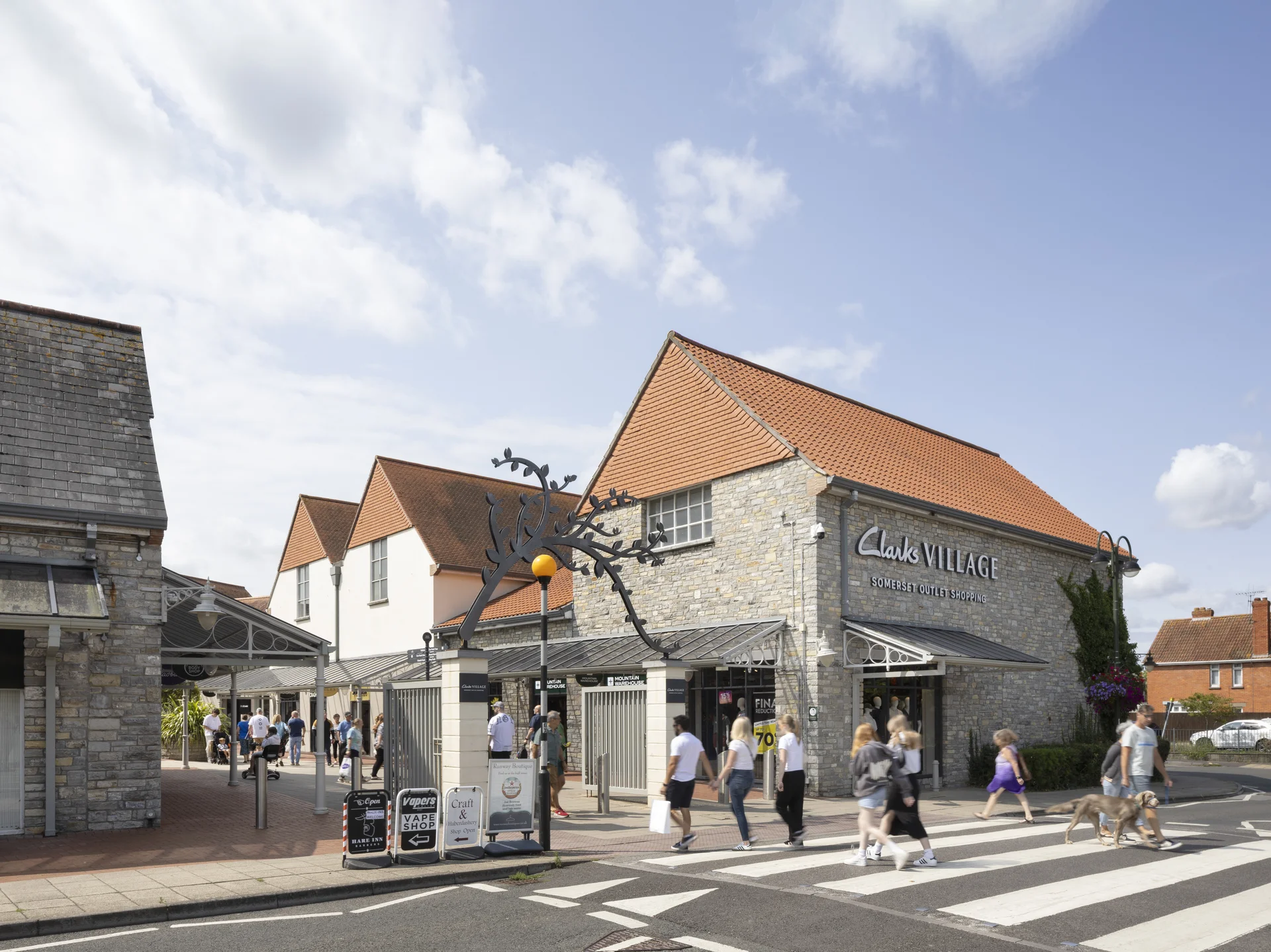 Exterior view of the main entrance to **Clarks Village** in Street, Somerset, featuring traditional stone buildings with orange tile roofs, and shoppers walking across the road, including a person with a dog.