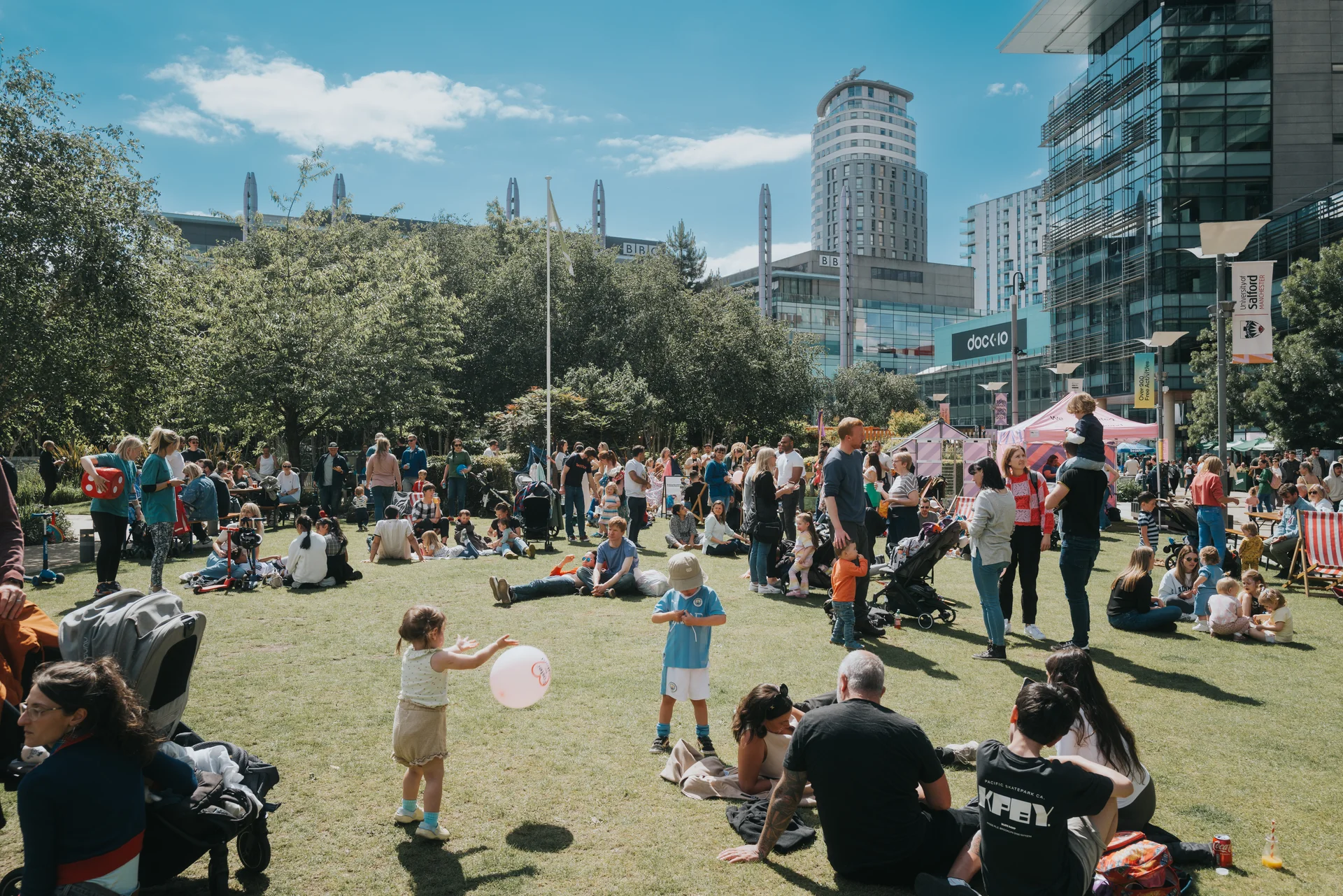 People enjoying a sunny day on a grassy lawn at MediaCityUK, with tall modern buildings and the Dock10 sign visible.