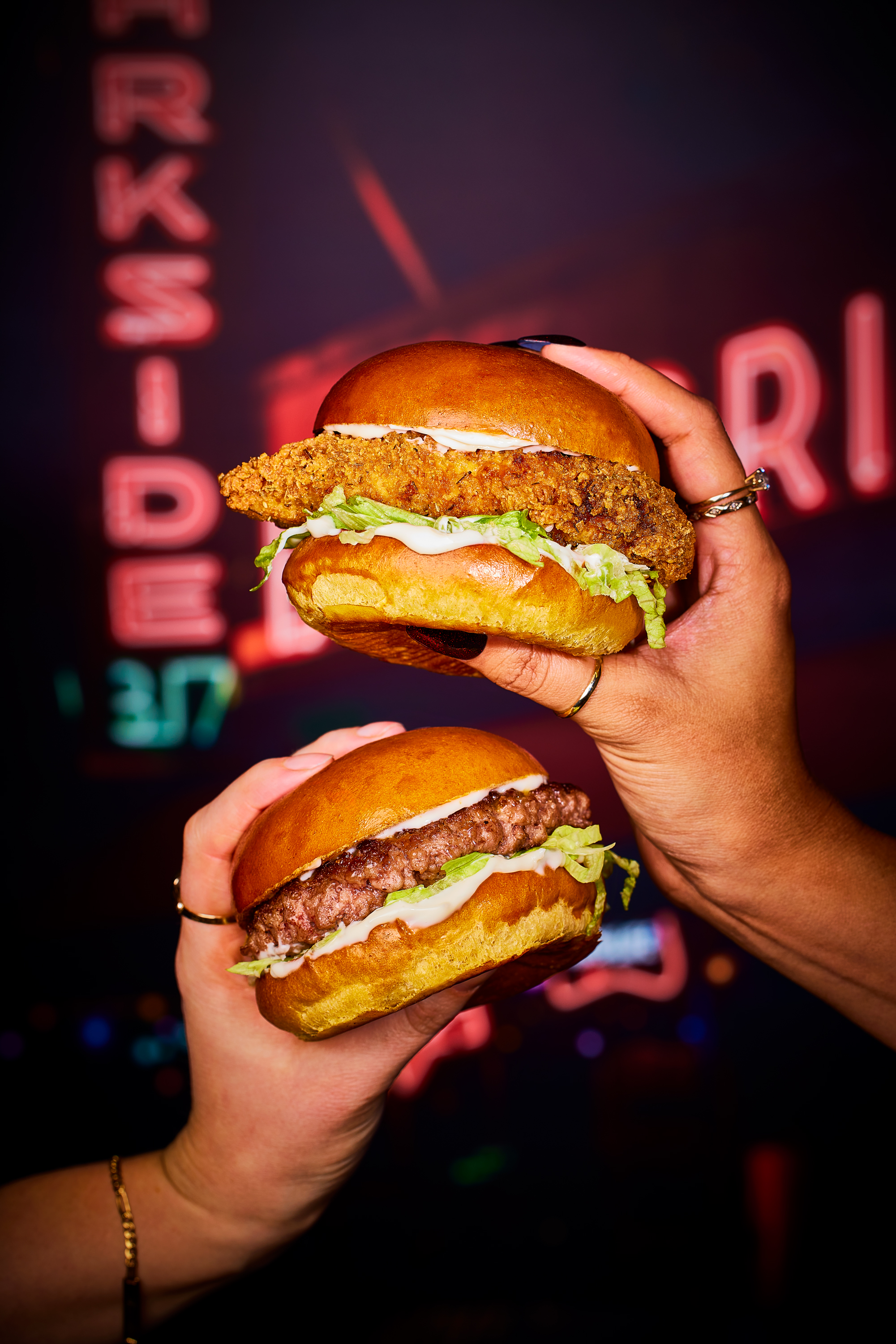 A close up shot of two burgers in two hands, one above the other, one chicken and one beef 