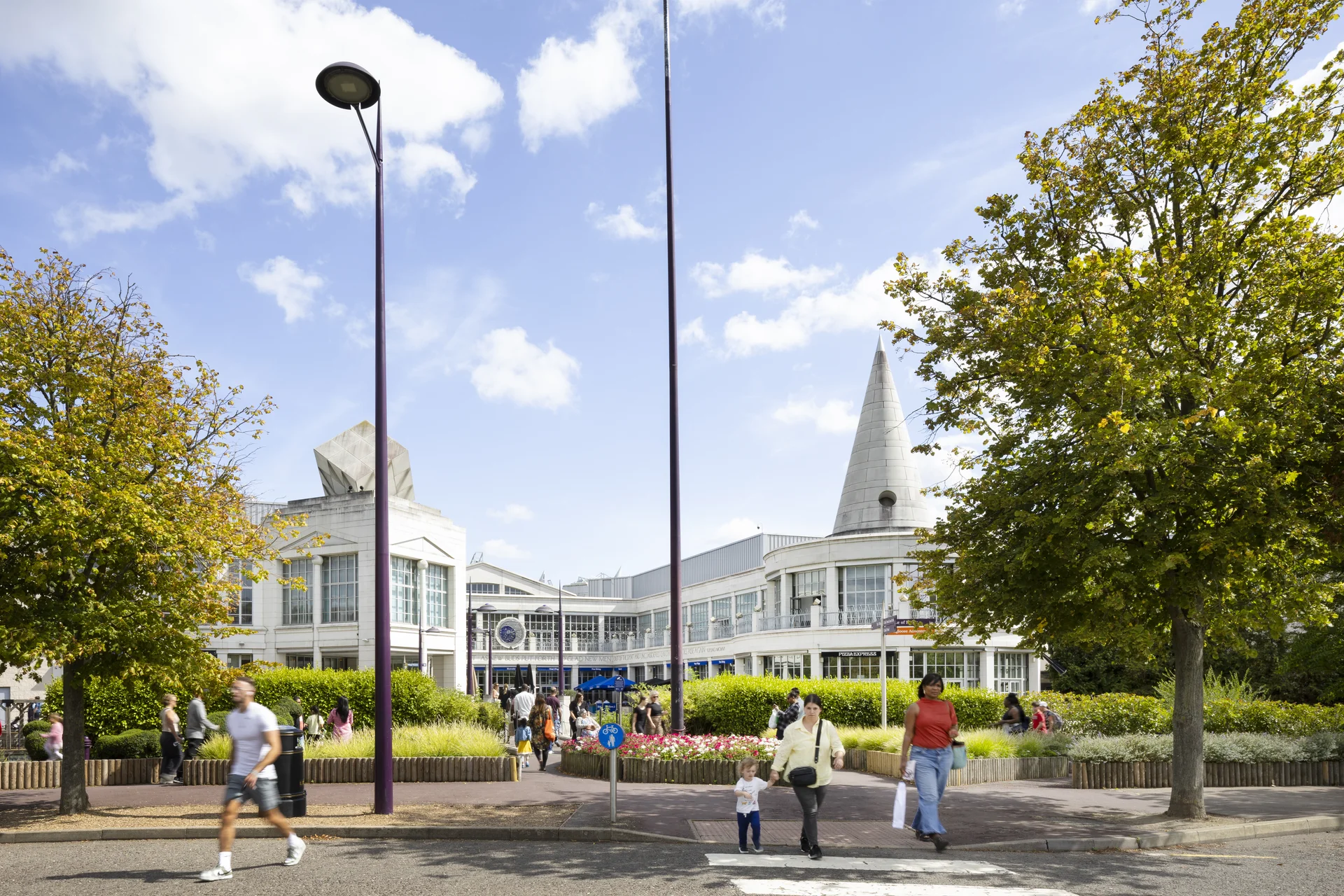 Exterior view of **Bluewater Shopping Centre** entrance on a sunny day, showcasing the distinctive white architectural features, including the conical roof, with trees and shoppers walking past flower beds.