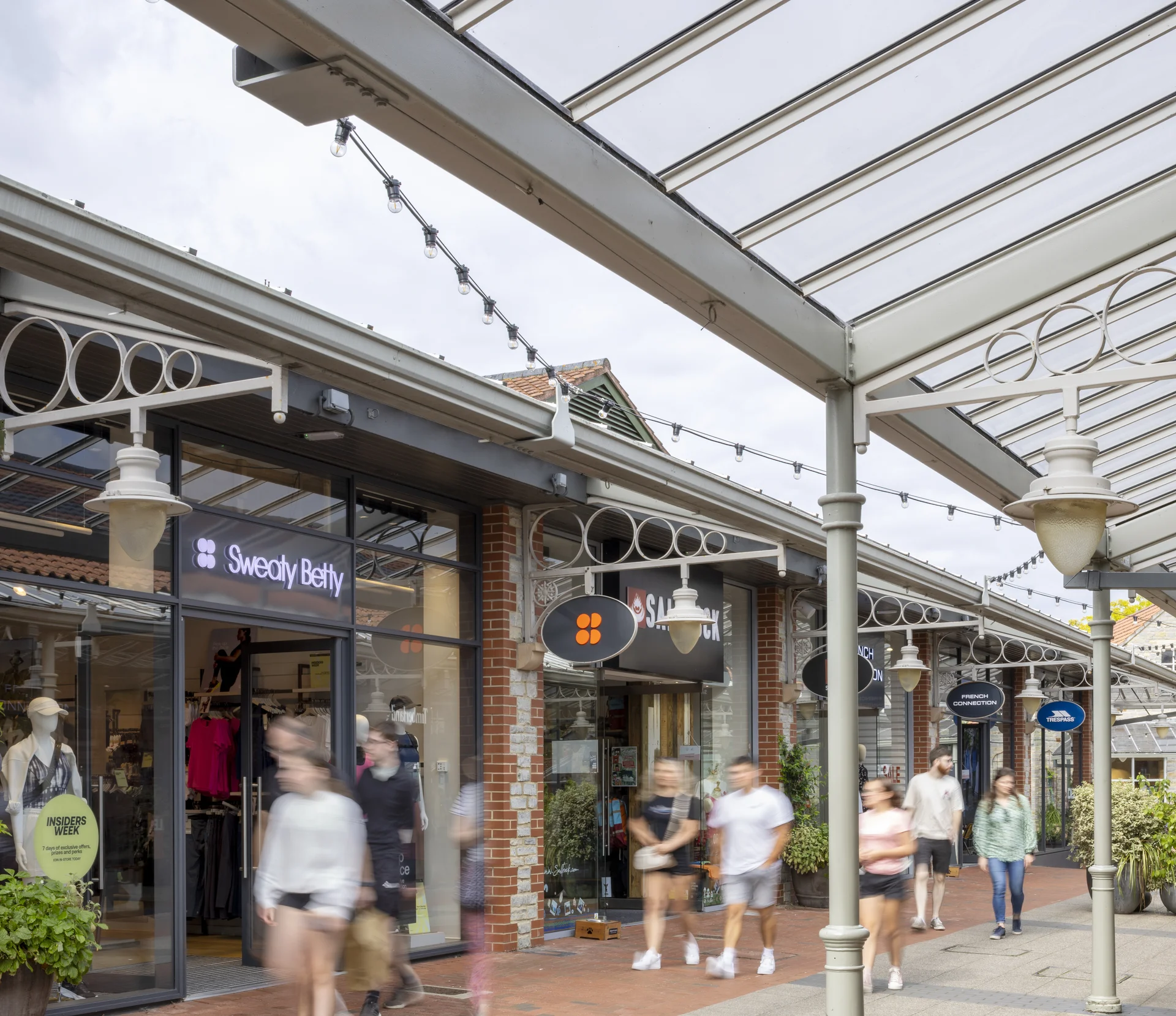 A covered outdoor walkway at **Clarks Village** outlet, featuring modern-traditional architecture, overhead string lights, and the storefronts for **Sweaty Betty** and **Salomon**, with blurred shoppers walking past.