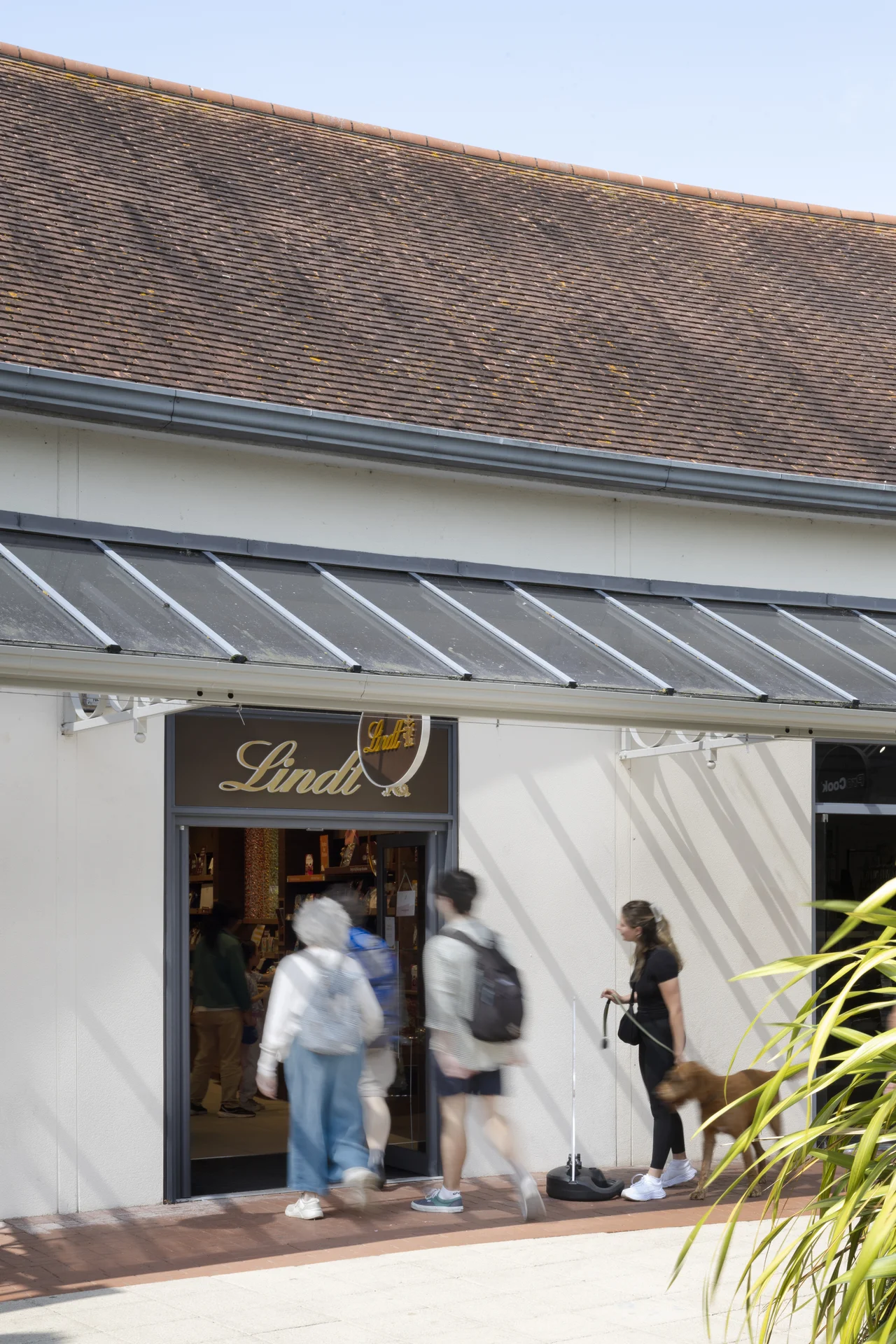 Shoppers entering the **Lindt** chocolate store at **Clarks Village**, with a minimalist white facade, a brown tiled roof, and a shopper standing near the entrance with a dog on a lead.