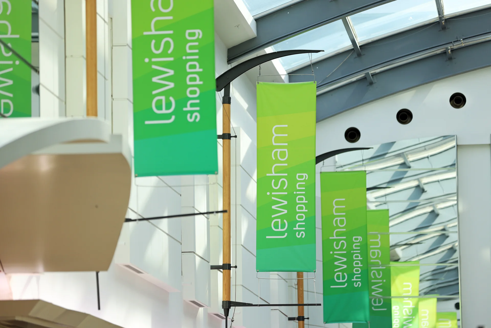 Green banners reading 'Lewisham shopping' hang from a modern building with glass and white panels, under a bright sky.